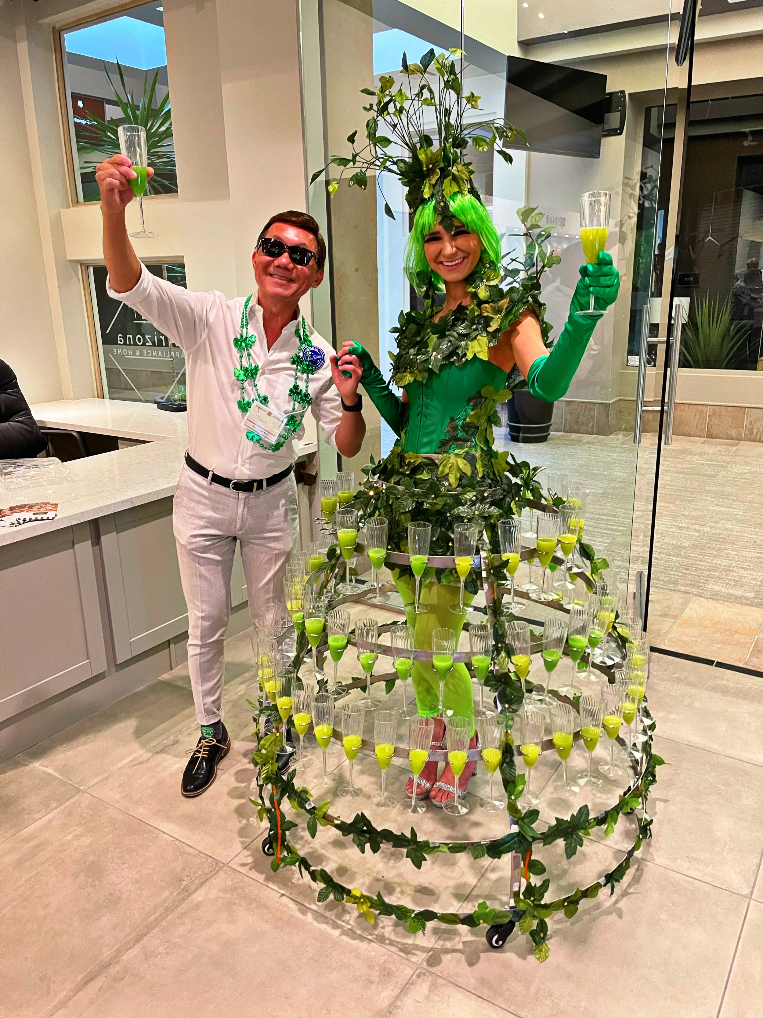 Person dressed in a green, nature-themed costume resembling a tree, standing next to a man in a white outfit with green accessories. The costume features an elaborate headpiece and is surrounded by a skirt-like structure holding multiple glasses fill