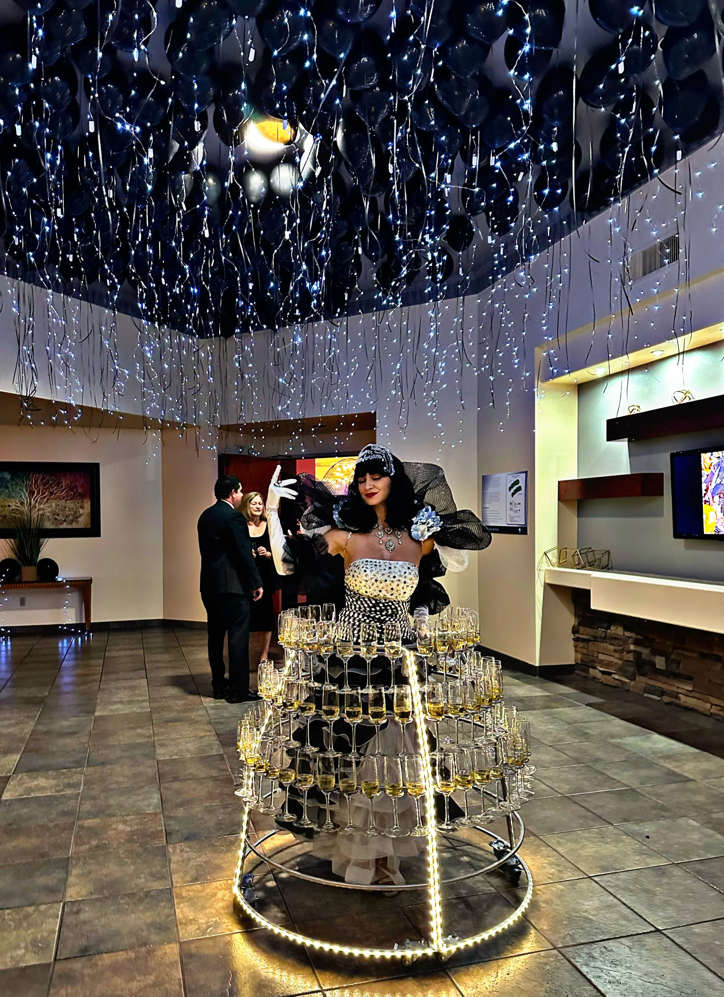 Woman in an illuminated dress holding multiple wine glasses, with balloon and string light decorations above, at an event with people in formal attire.