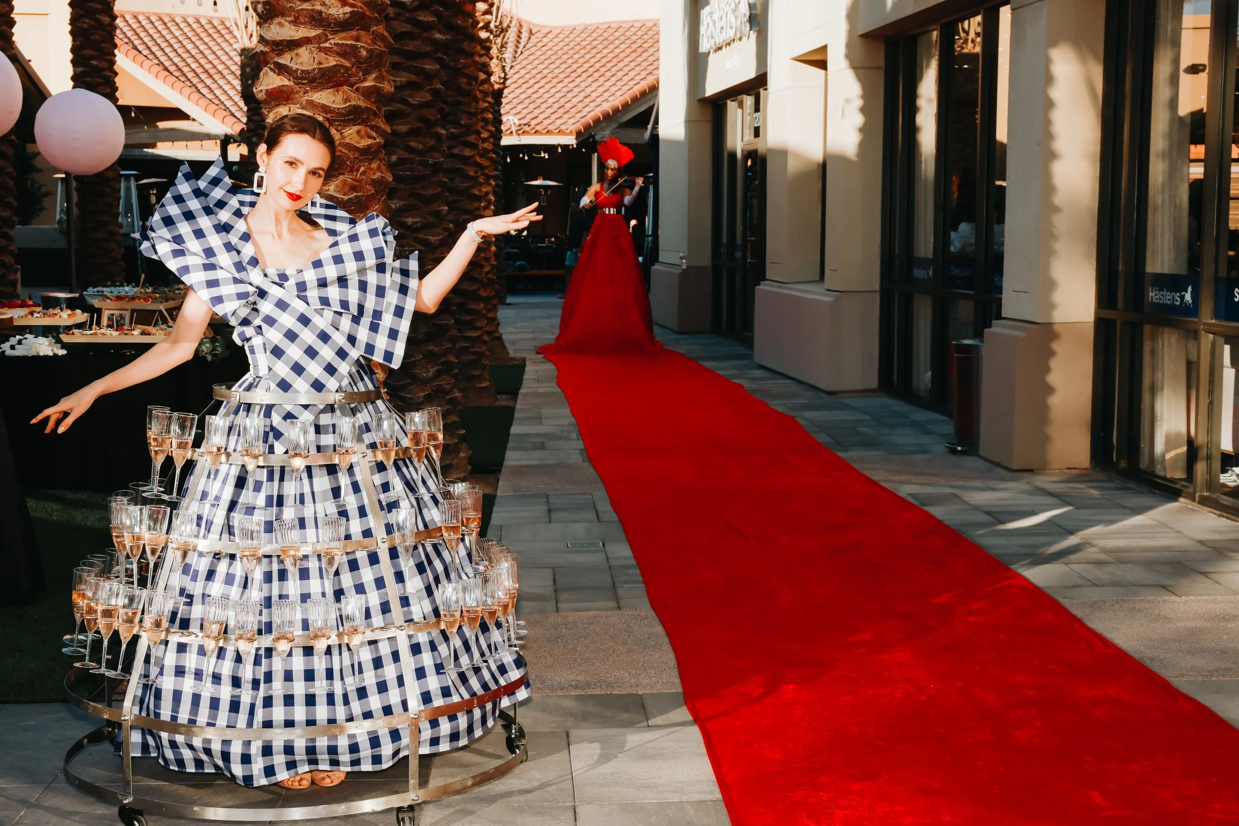 Woman in a blue and white checkered dress holding champagne glasses, standing near a red carpet with a violinist in the background.