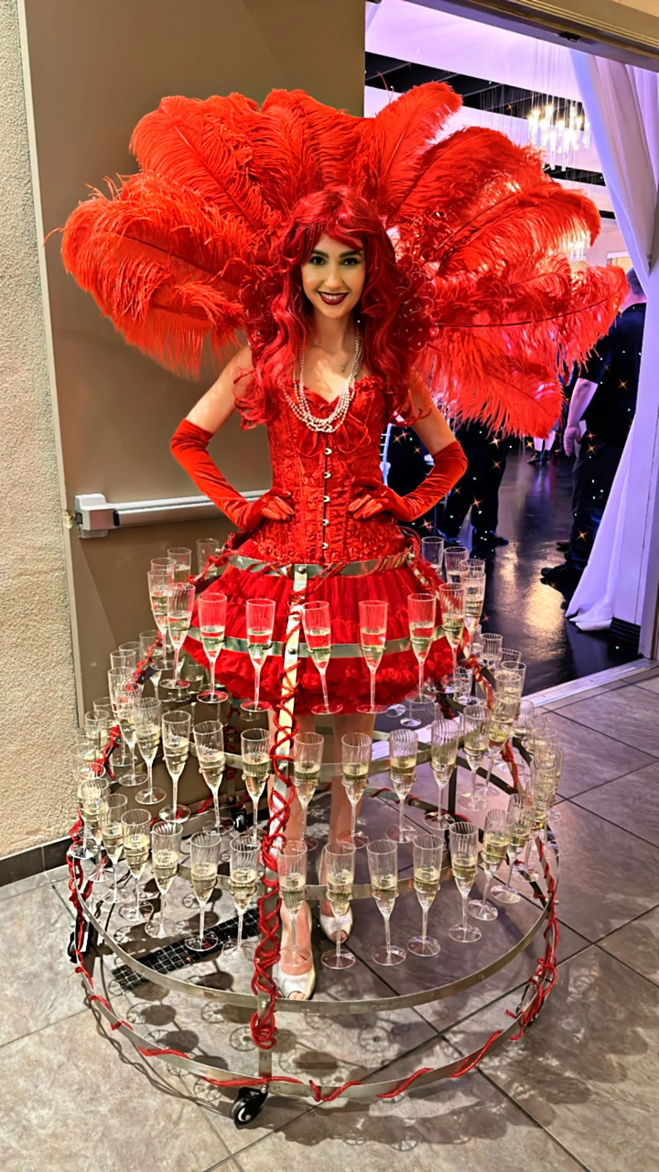 Woman in elaborate red costume with feather headdress, surrounded by champagne glasses on a mobile rack.