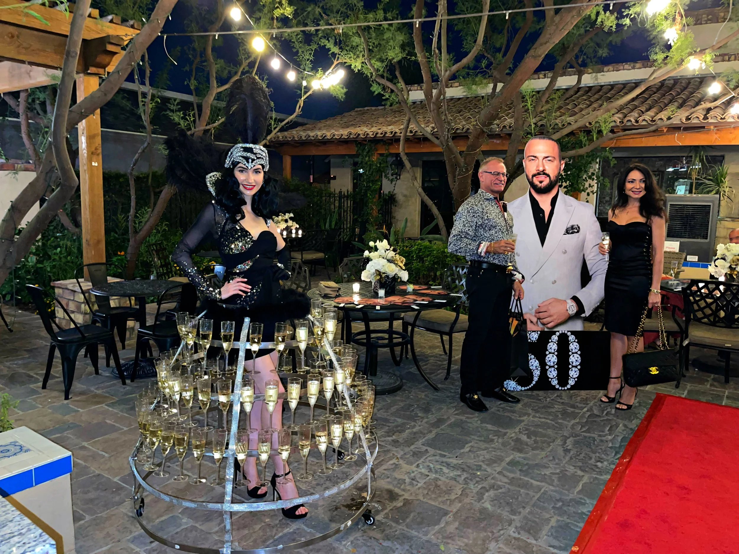 Outdoor party scene with a woman dressed in a black costume adorned with champagne glasses, standing near two people posing by a large cut-out photo. Decor includes a table with white flowers and strings of lights hanging above.