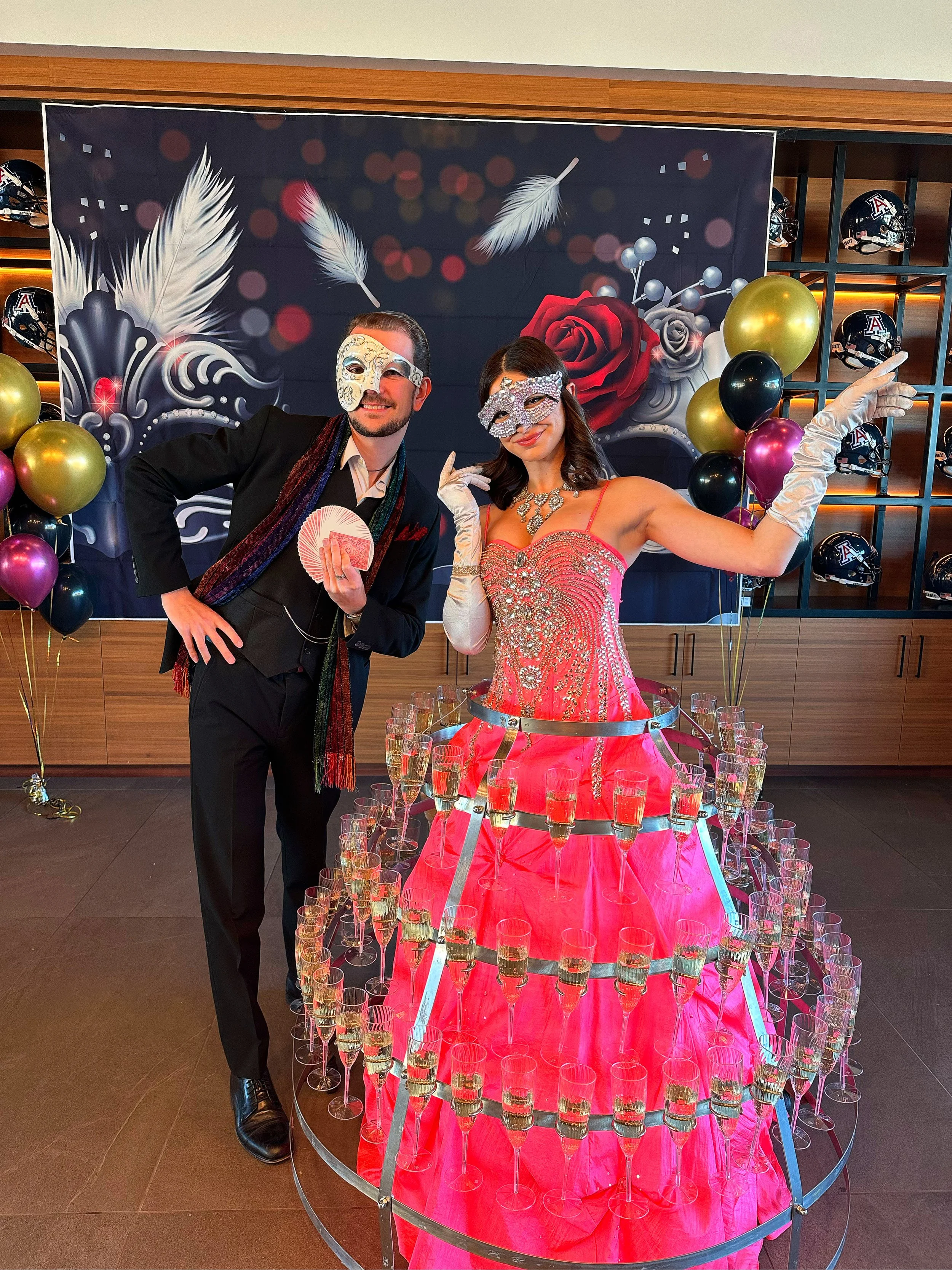 Two people wearing masquerade masks, standing next to a champagne dress display. The woman is in a pink dress adorned with glasses of champagne, while the man holds playing cards. Decorative background with feathers, roses, and balloons.