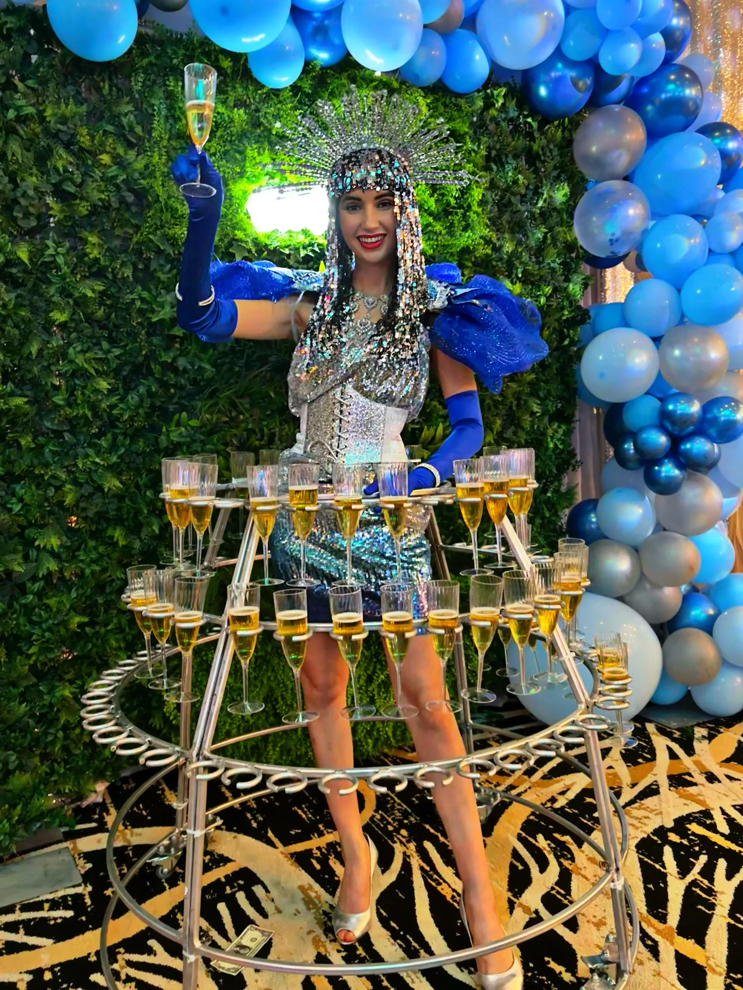Person dressed in a sparkling blue and silver costume, surrounded by a circular champagne glass holder filled with glasses, standing in front of a green and blue balloon backdrop.
