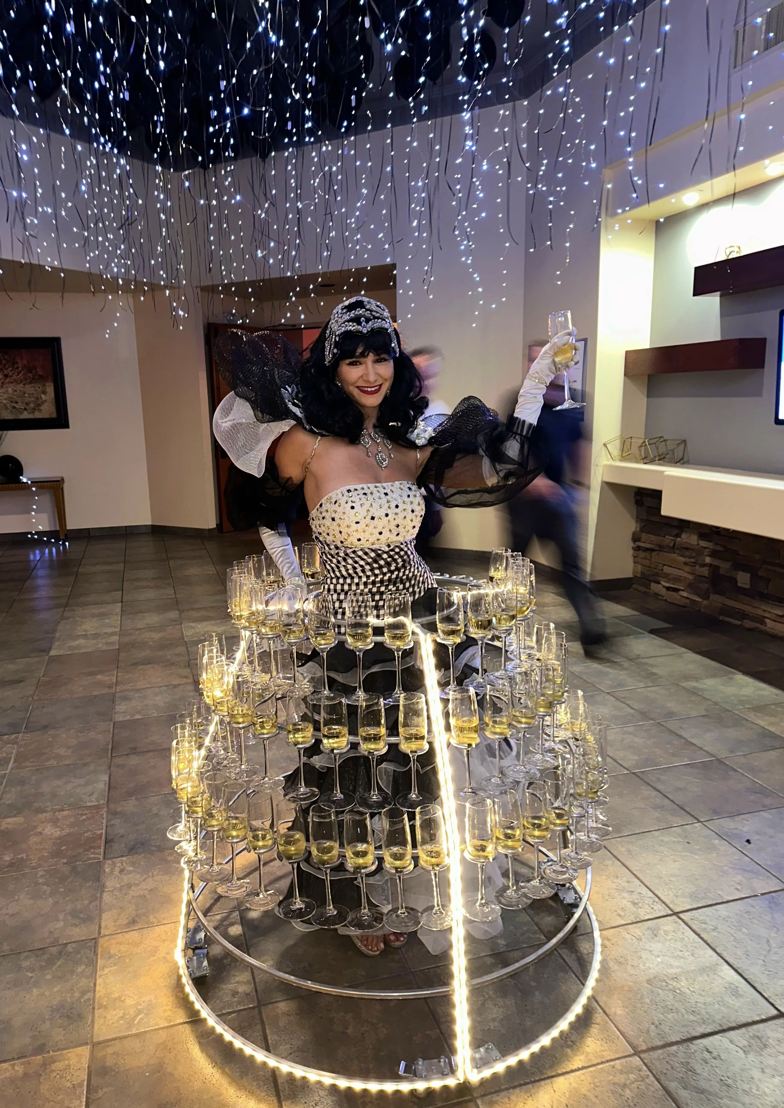 A woman dressed in an elaborate outfit stands within a circular wine glass display, holding a glass of champagne. The display is adorned with string lights and surrounded by hanging decorations.