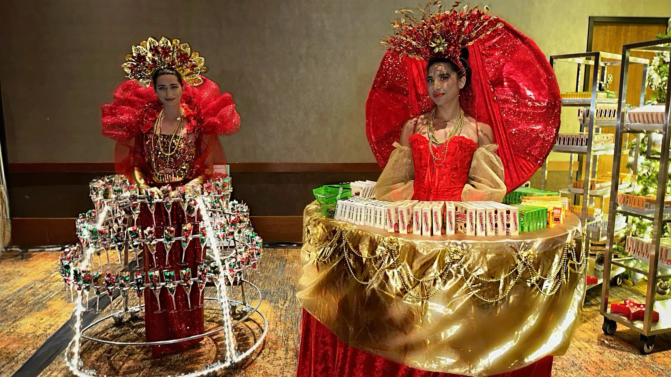 Two people dressed in elaborate red and gold costumes, each with a wearable table displaying various items, standing indoors.