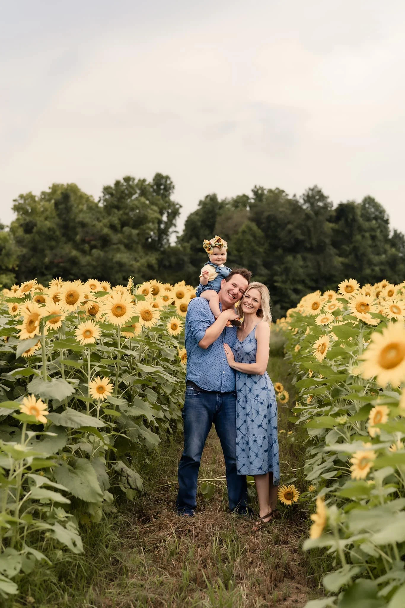 a family of a mom, dad and baby are in a field of sunflowers. The baby is on dad's shoulders and mom and dad are snuggled in together. The family is smiling at the camera.