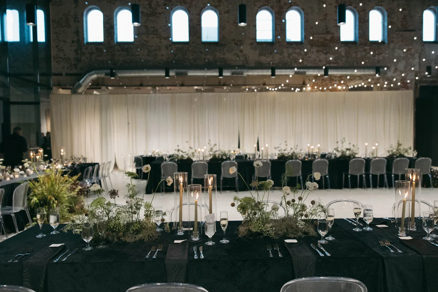Elegant banquet hall decorated for a formal event with long tables, floral centerpieces, candles, and string lights, set against an exposed brick wall with high arched windows.