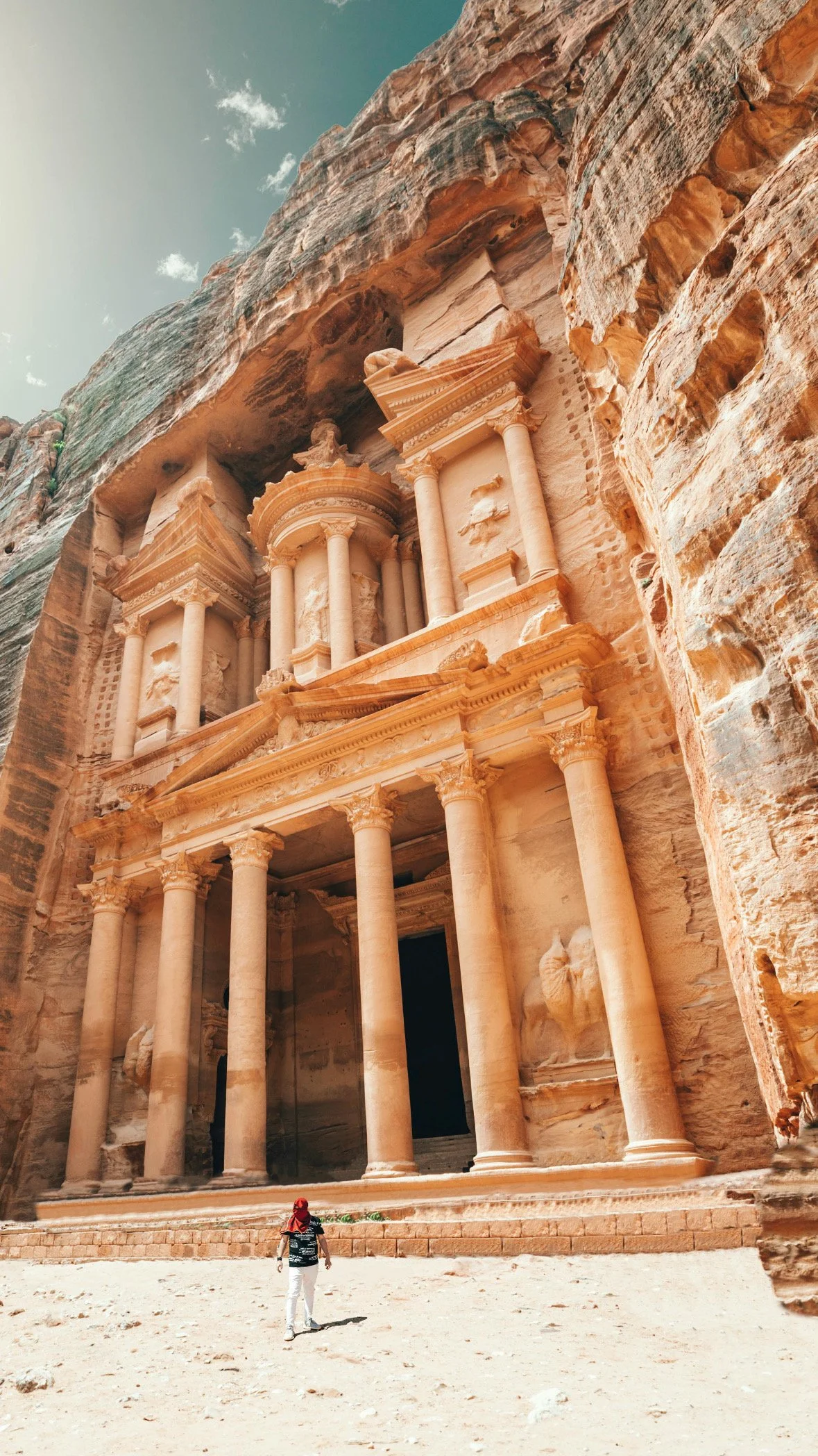 The image shows the famous ancient rock-cut city of Petra in Jordan, featuring the intricate facades of tombs and structures carved into the pink sandstone cliffs. A person wearing a red headscarf and black top stands in front of the grand face of the Treasury building.