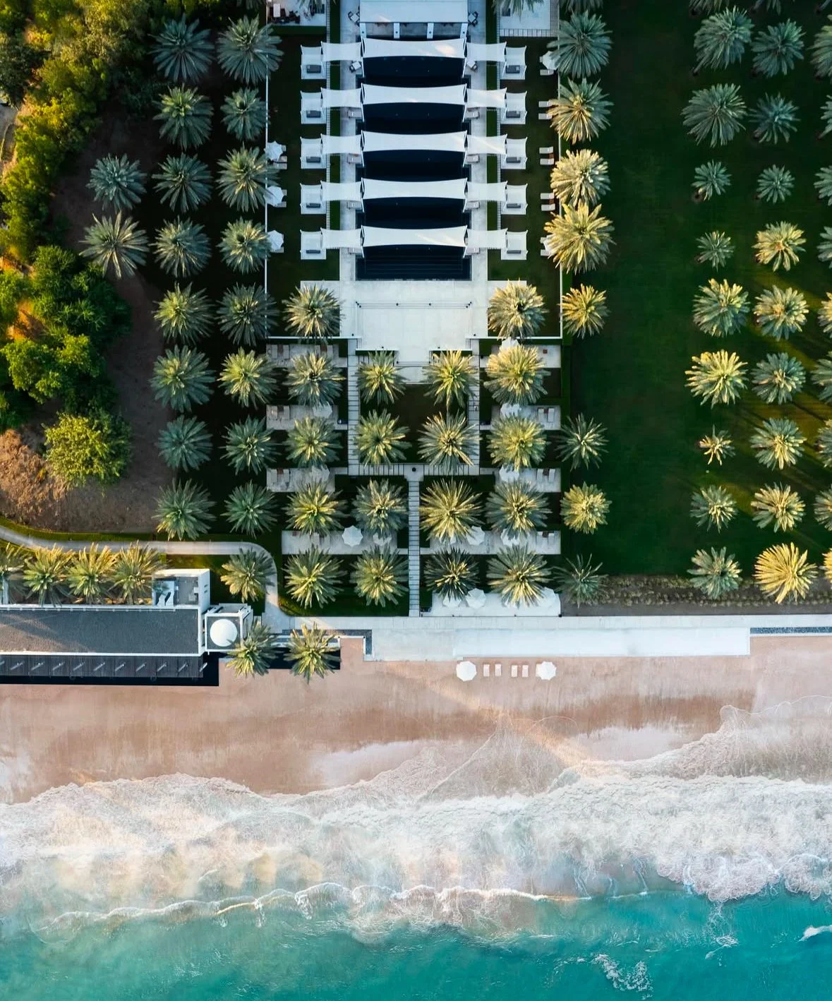 An aerial view of a tropical beachside resort in Bali featuring a row of cabanas with white roofs, surrounded by numerous palm trees, with a sandy beach and ocean waves at the bottom of the image.