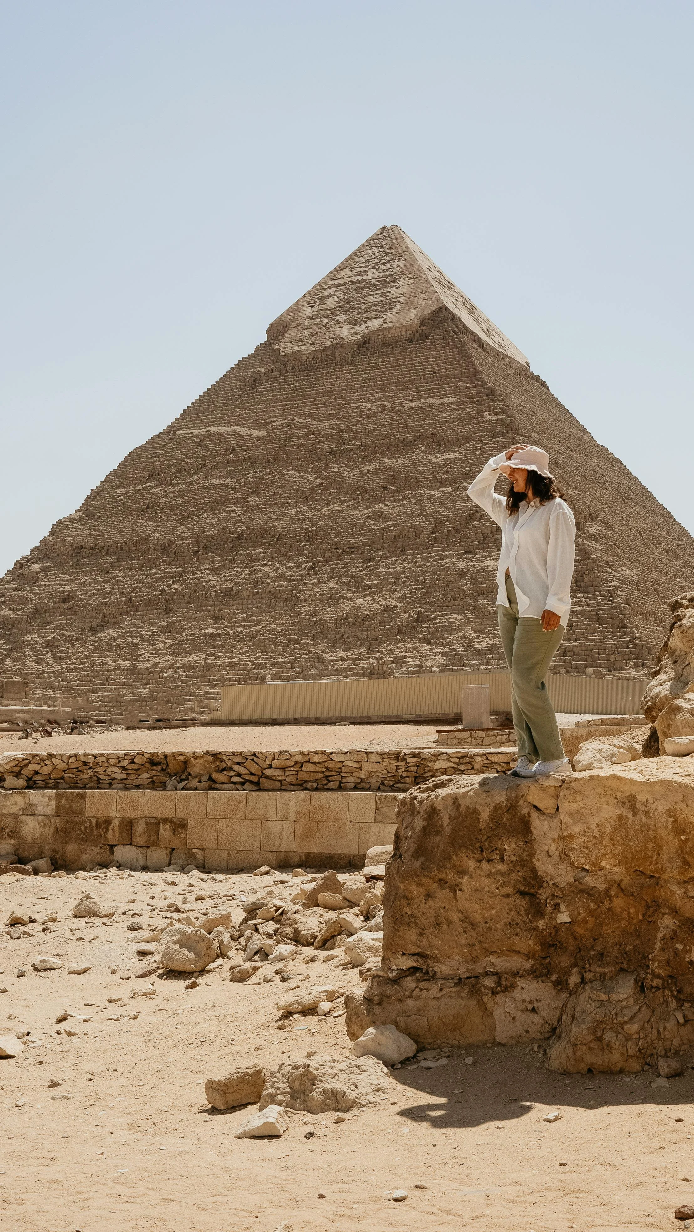 Woman standing on a large rock in front of the Great Pyramid of Giza, Egypt wearing a white shirt, beige pants, and a sun hat, shielding her eyes from the sun.