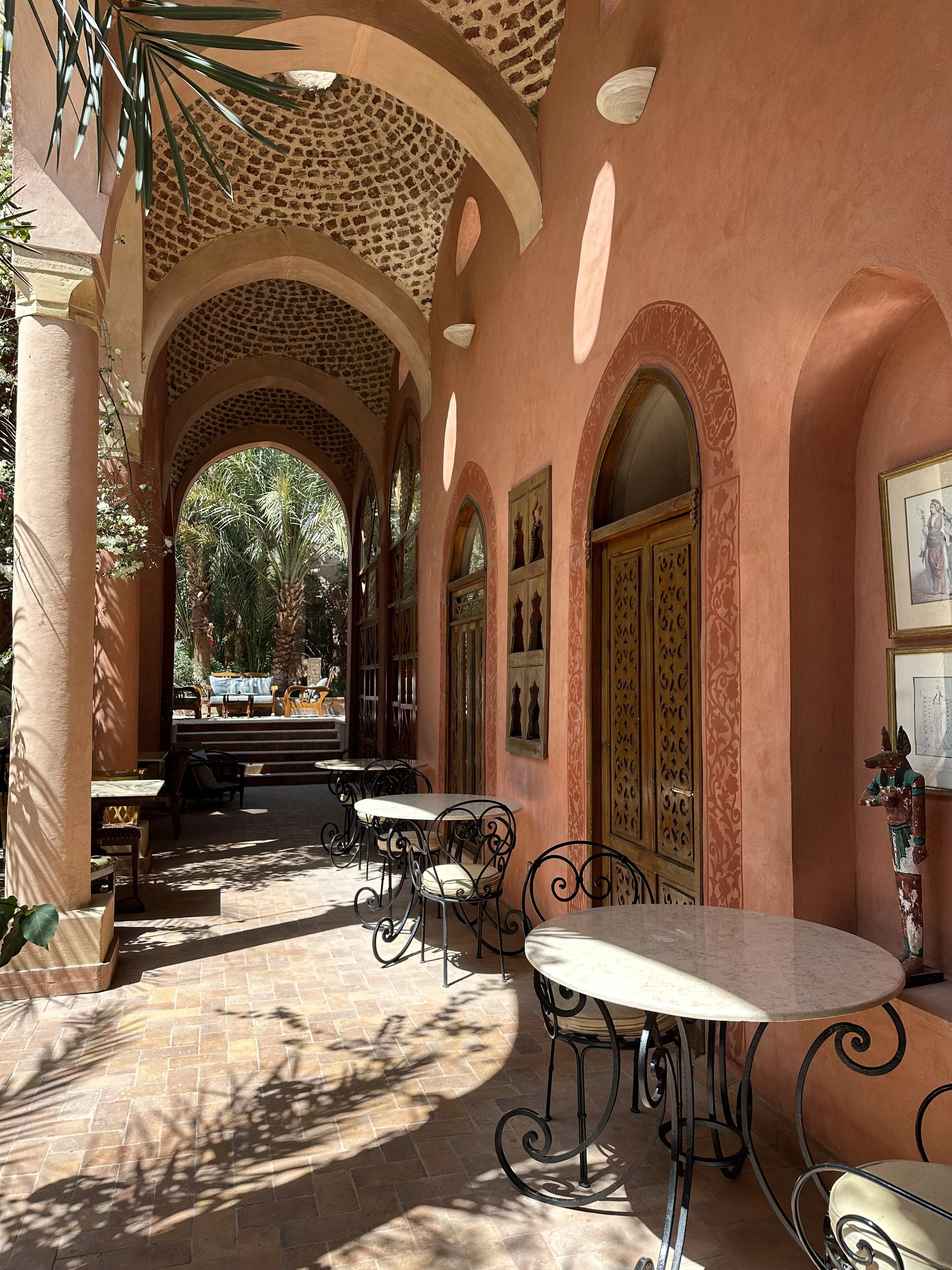 Sunlit terrace with pink stucco walls, arched windows and doorways, decorative artwork, and wrought iron round tables and chairs with white cushions, adjacent to lush palm trees.