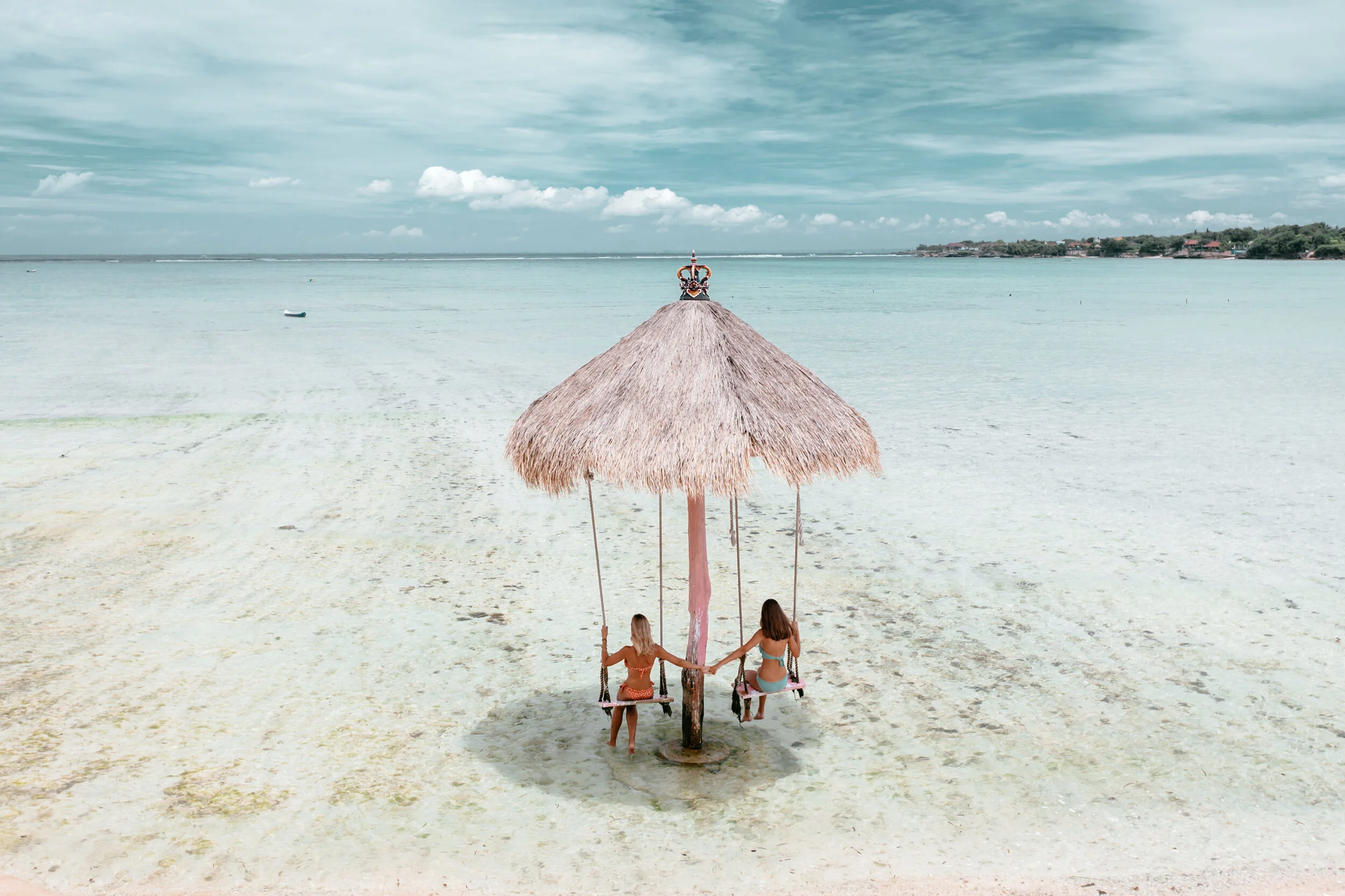 Two women sitting on swings under a thatched umbrella in Bali on a sandy beach with calm ocean and cloudy sky in the background.