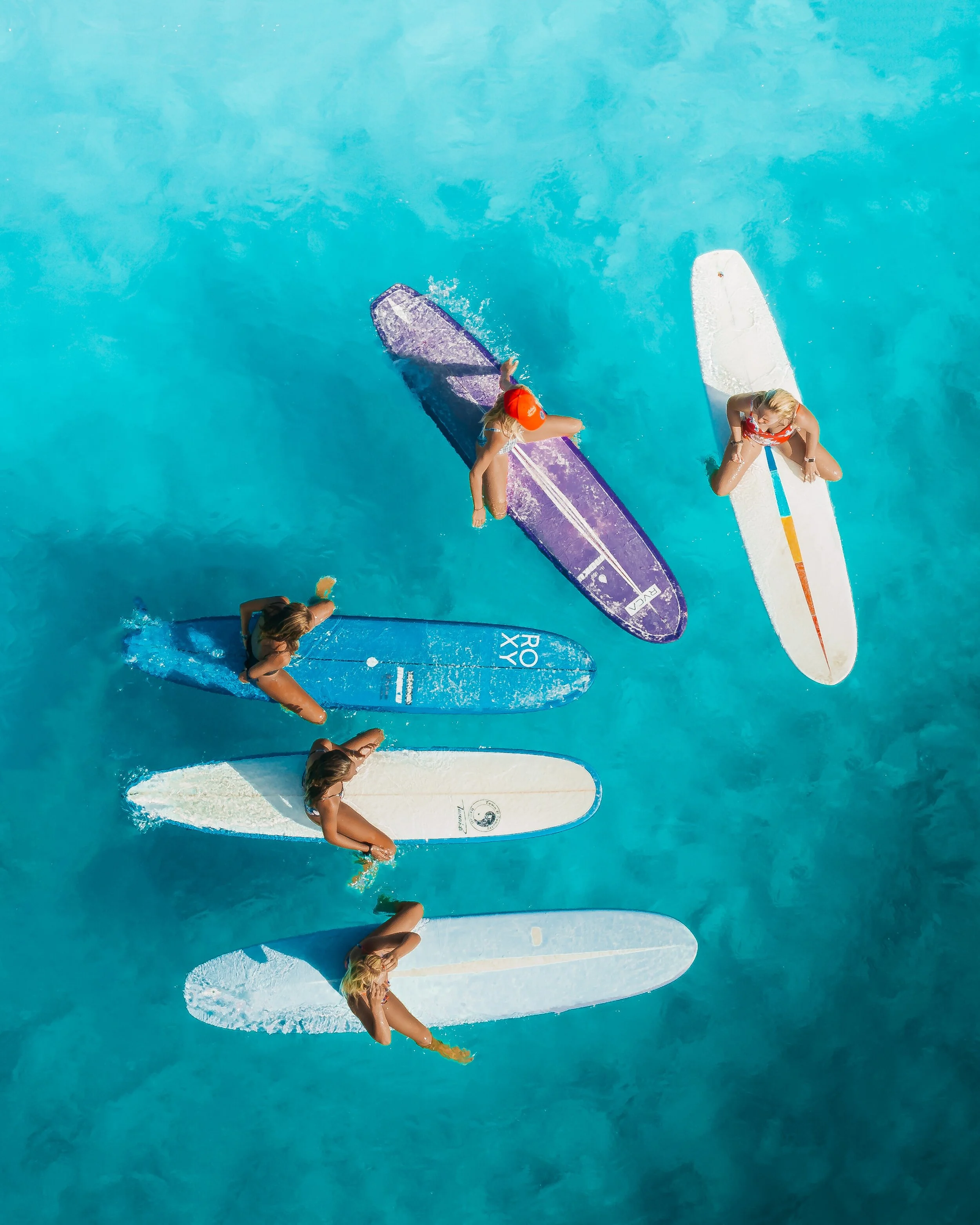 Five women are floating on stand-up paddleboards in turquoise water, viewed from above in Bali.