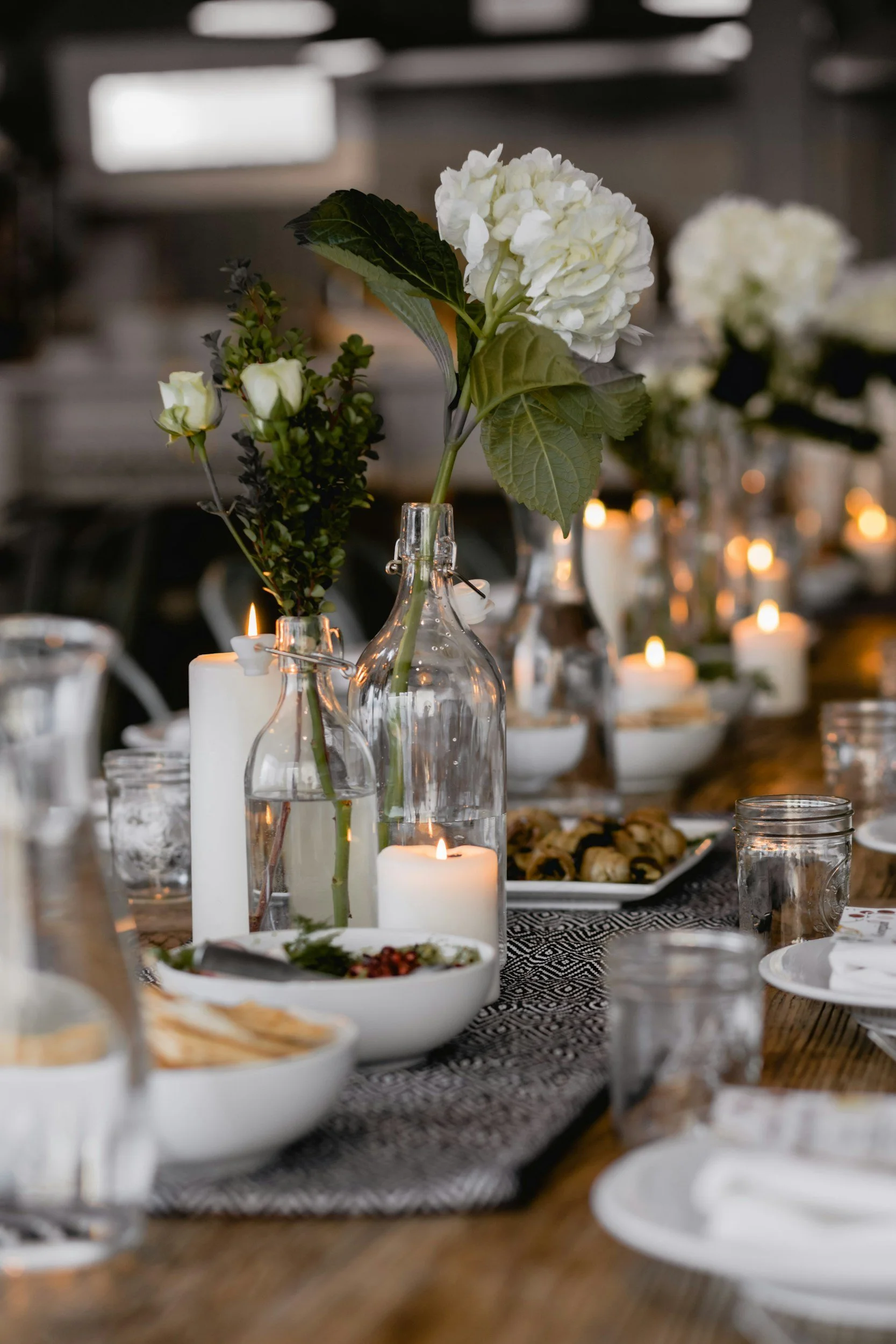 A styled dining table with candles and glass bottles holding white flowers and greenery.