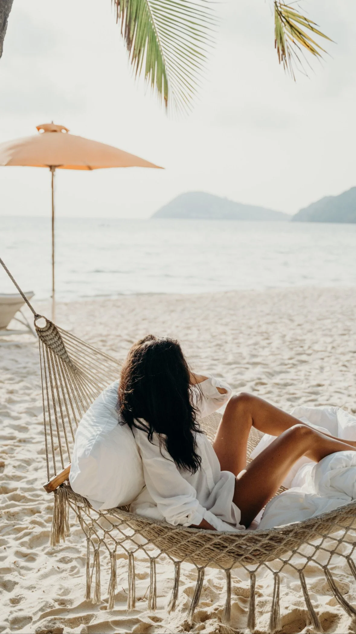Woman relaxing in a hammock on a sandy beach in Bali with an umbrella and ocean in the background.