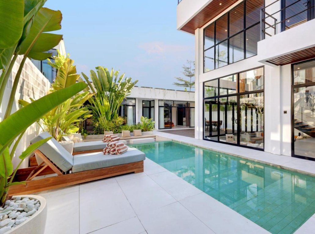 Modern house with large glass windows, a small swimming pool, and outdoor seating area with lounge chairs and tropical plants.