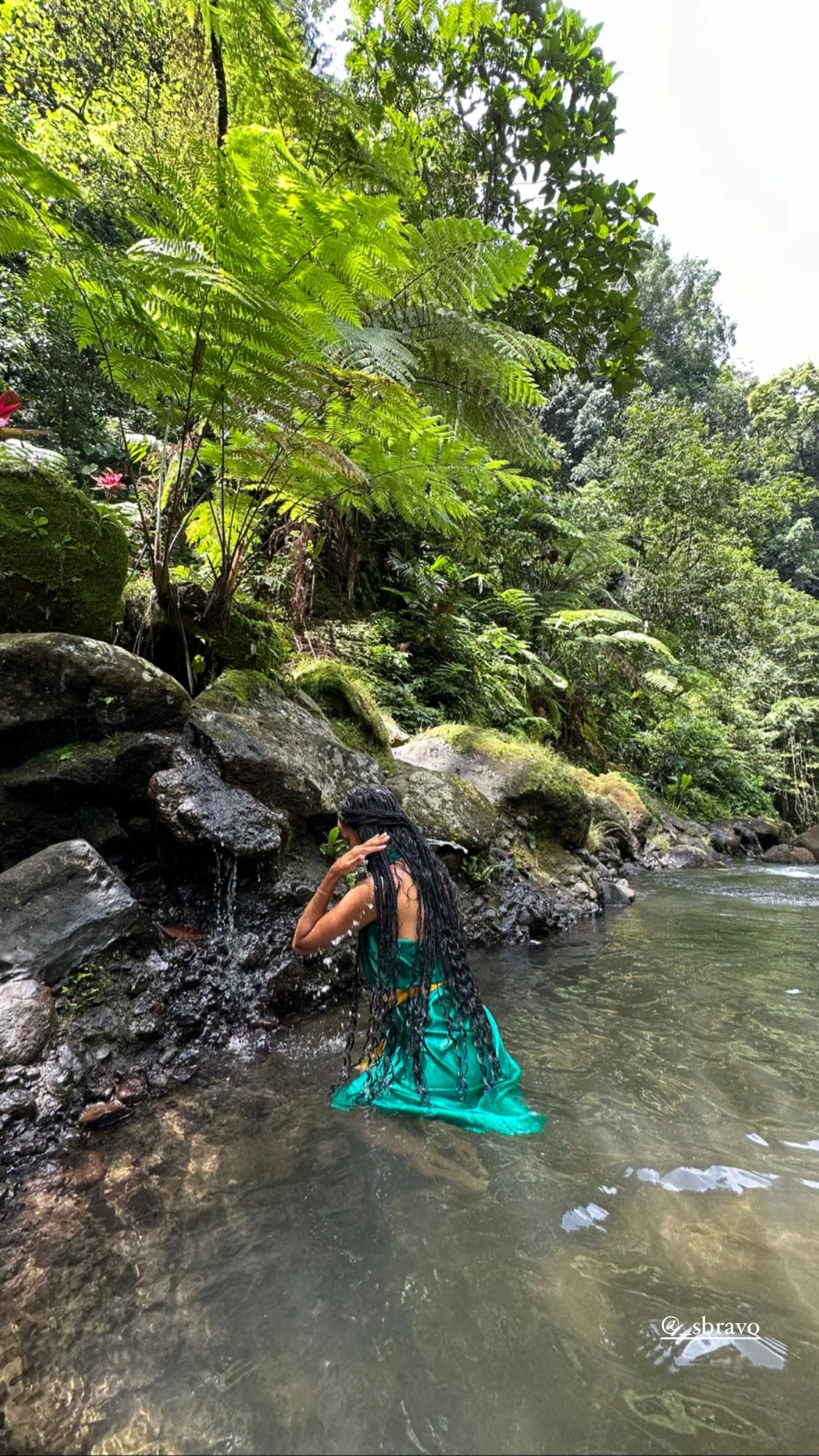 A woman with long braided hair wearing a green dress in a river, surrounded by lush green trees and rocks in Bali.