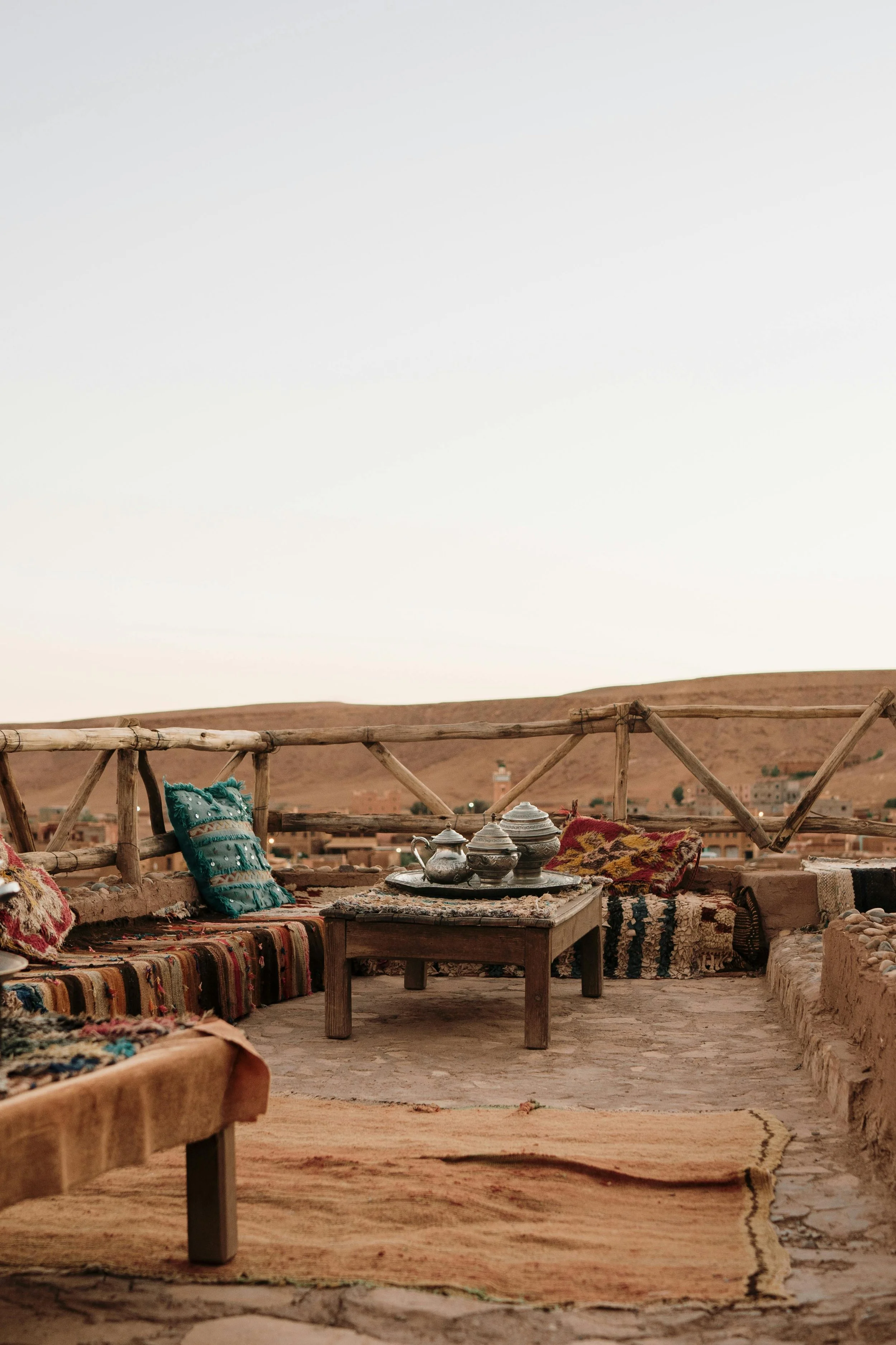 An outdoor seating area with traditional rugs and cushions on a rooftop, overlooking a desert landscape with distant buildings and a clear sky.
