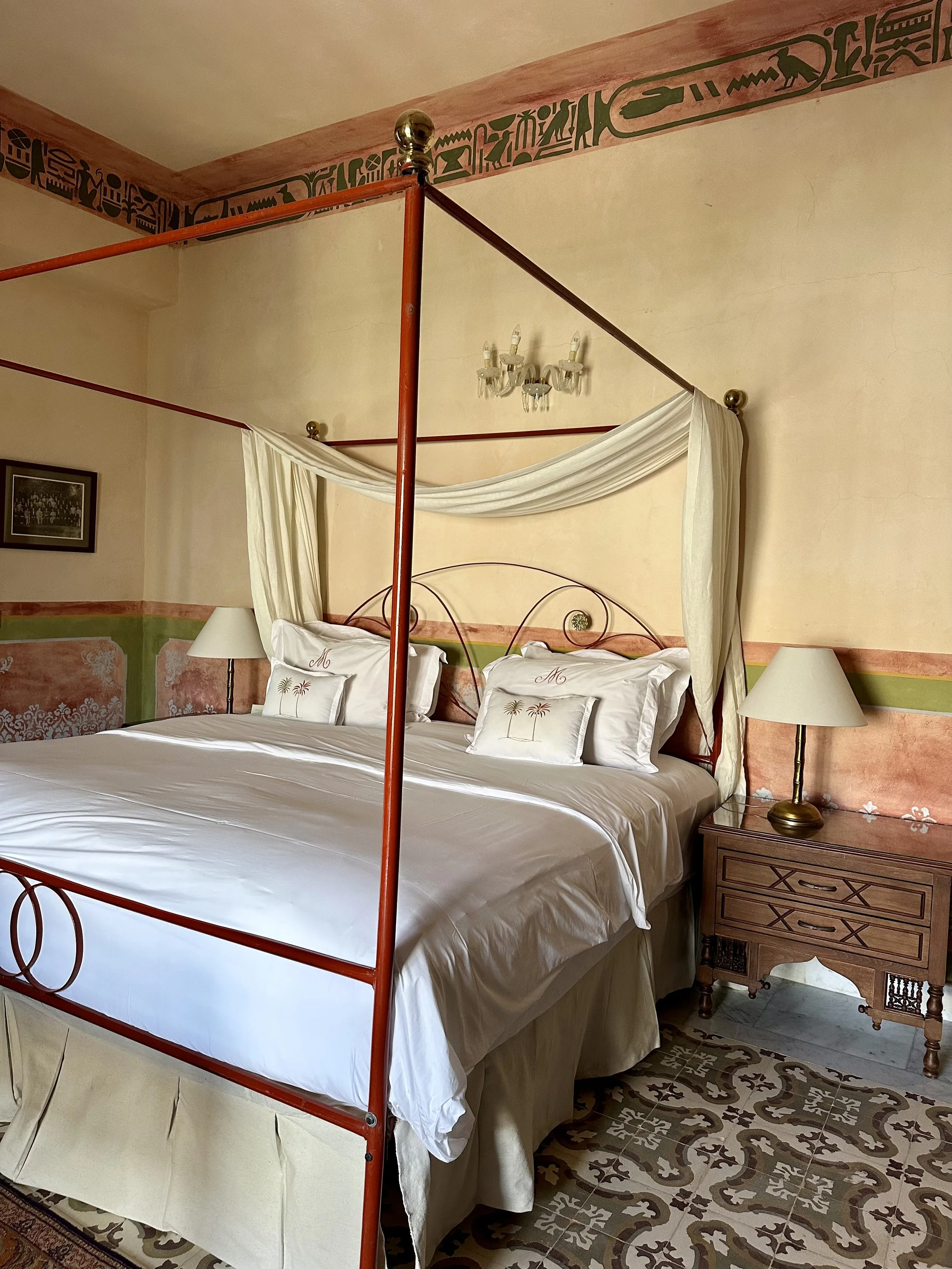 A bedroom with a four-poster bed with white sheets and pillows, surrounded by beige and pinkish walls, with a wooden nightstand and lamps on each side.
