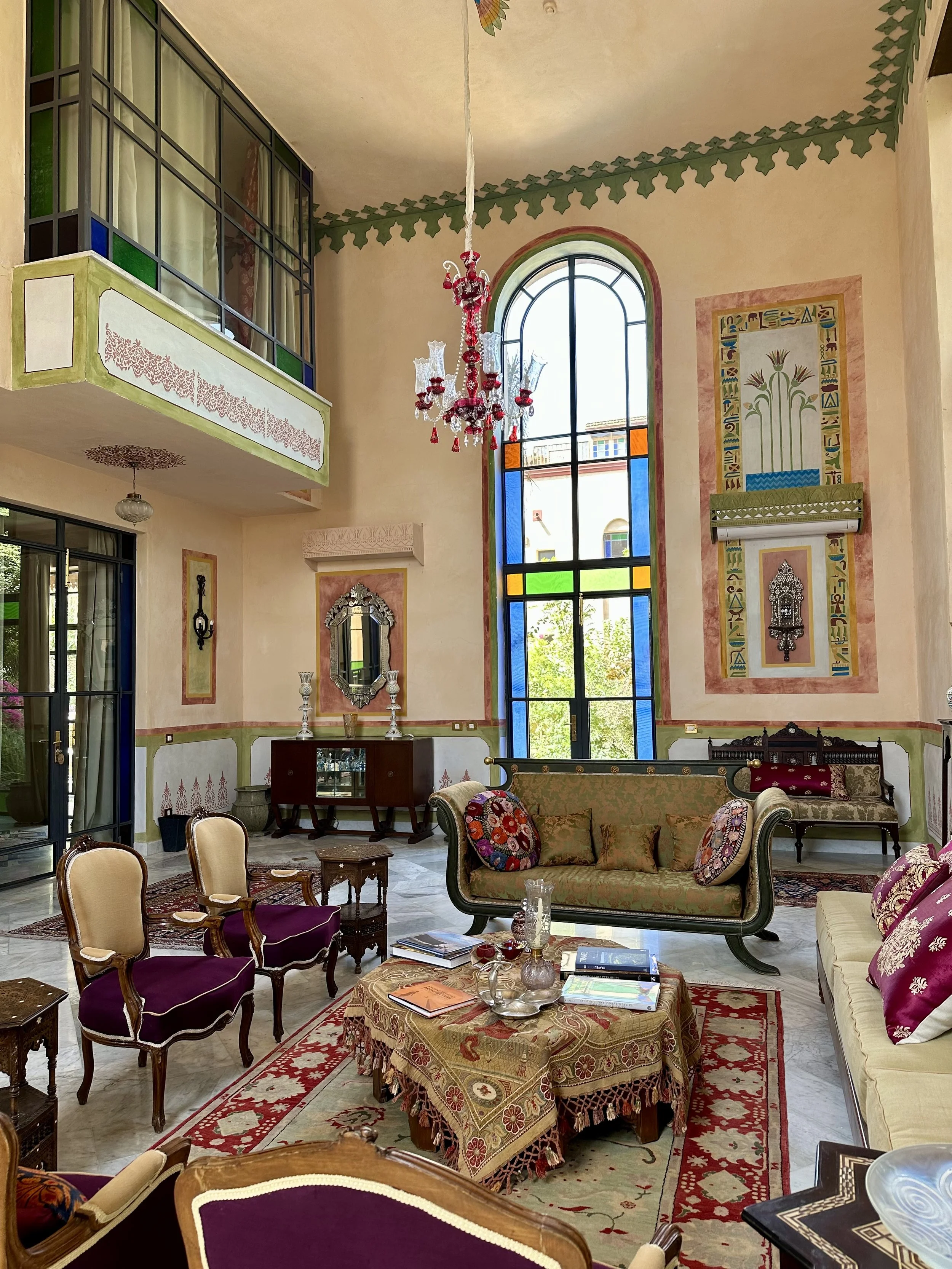 Interior of a vintage-style living room with tall colorful stained glass window, ornate chandelier, antique furniture, Persian rugs, and decorative wall art.
