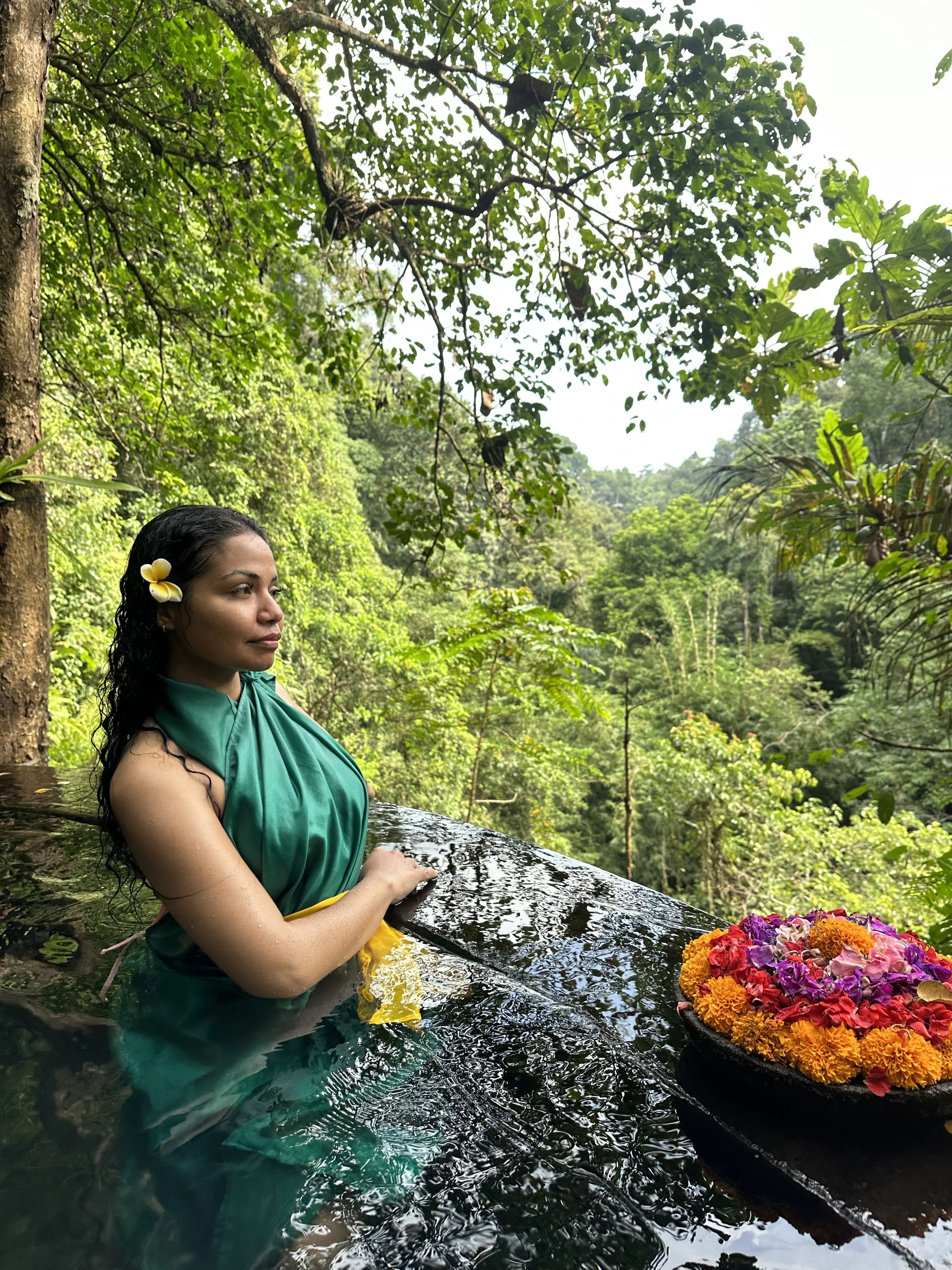 A woman with a yellow flower in her dark, curly hair, dressed in a green sarong, is in a natural outdoor hot spring or pool with lush green trees and forest in the background. She is in profile with her eyes closed, holding her arms on the edge of the pool. A black bowl of colorful flowers is on the edge of the pool.