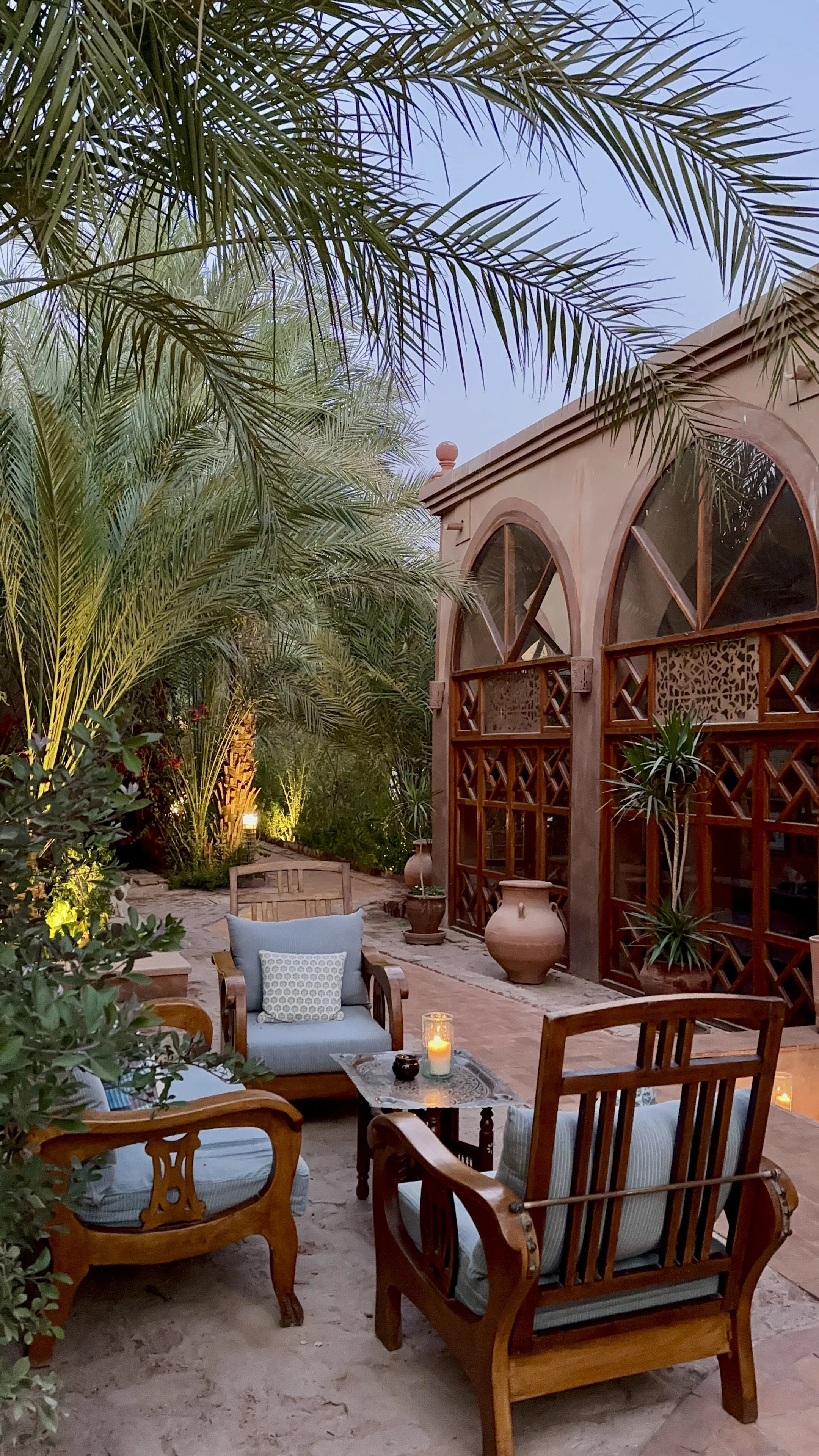 An outdoor patio with wooden furniture, potted plants, and lit candles, surrounded by lush green palm trees and a building with arched windows at dusk.