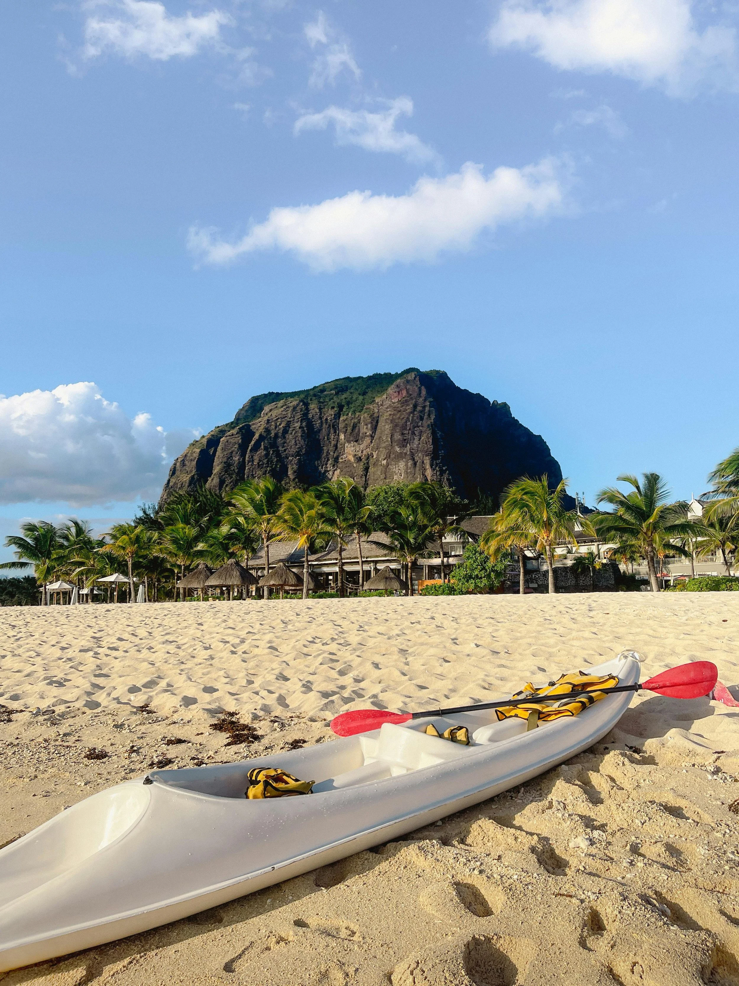 A white kayak with red paddles resting on a sandy beach, with a line of palm trees, beach huts, and a large mountain in the background under a partly cloudy sky.