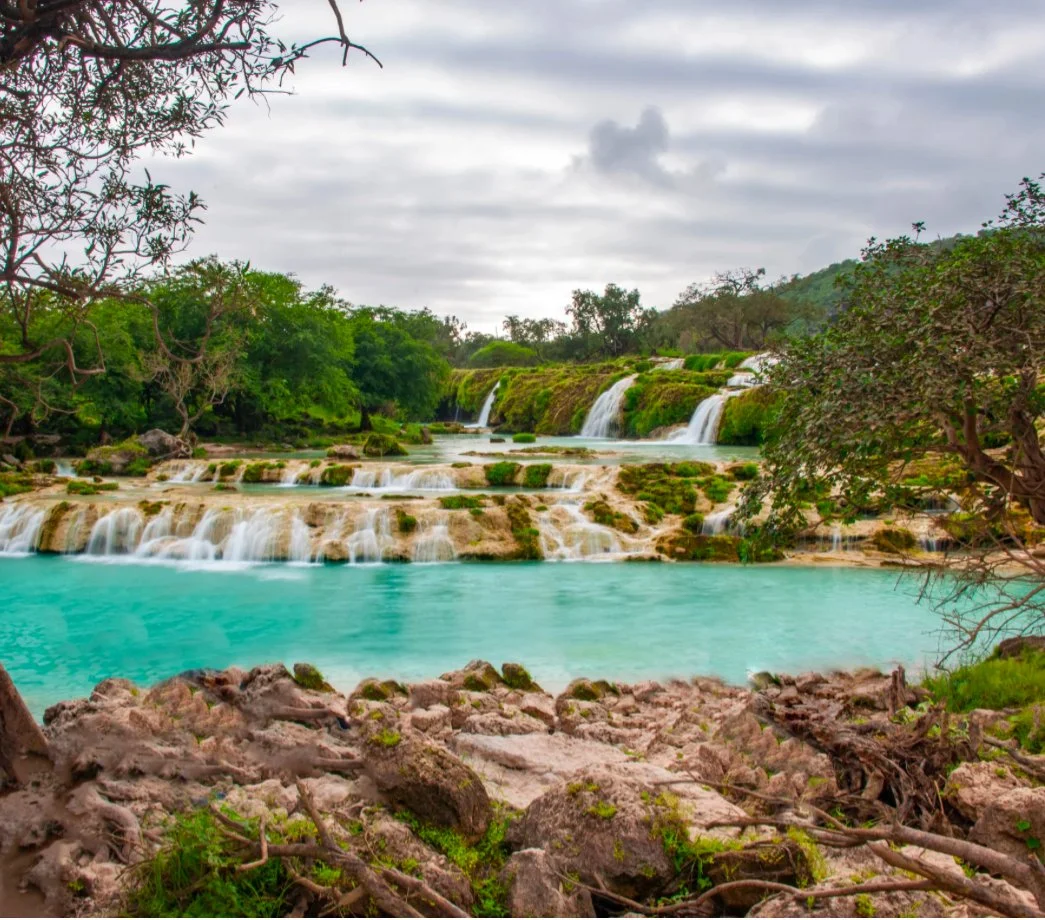 A serene scene of waterfalls cascading into a turquoise river, surrounded by lush green trees and rocks, with a cloudy sky overhead in Bali.
