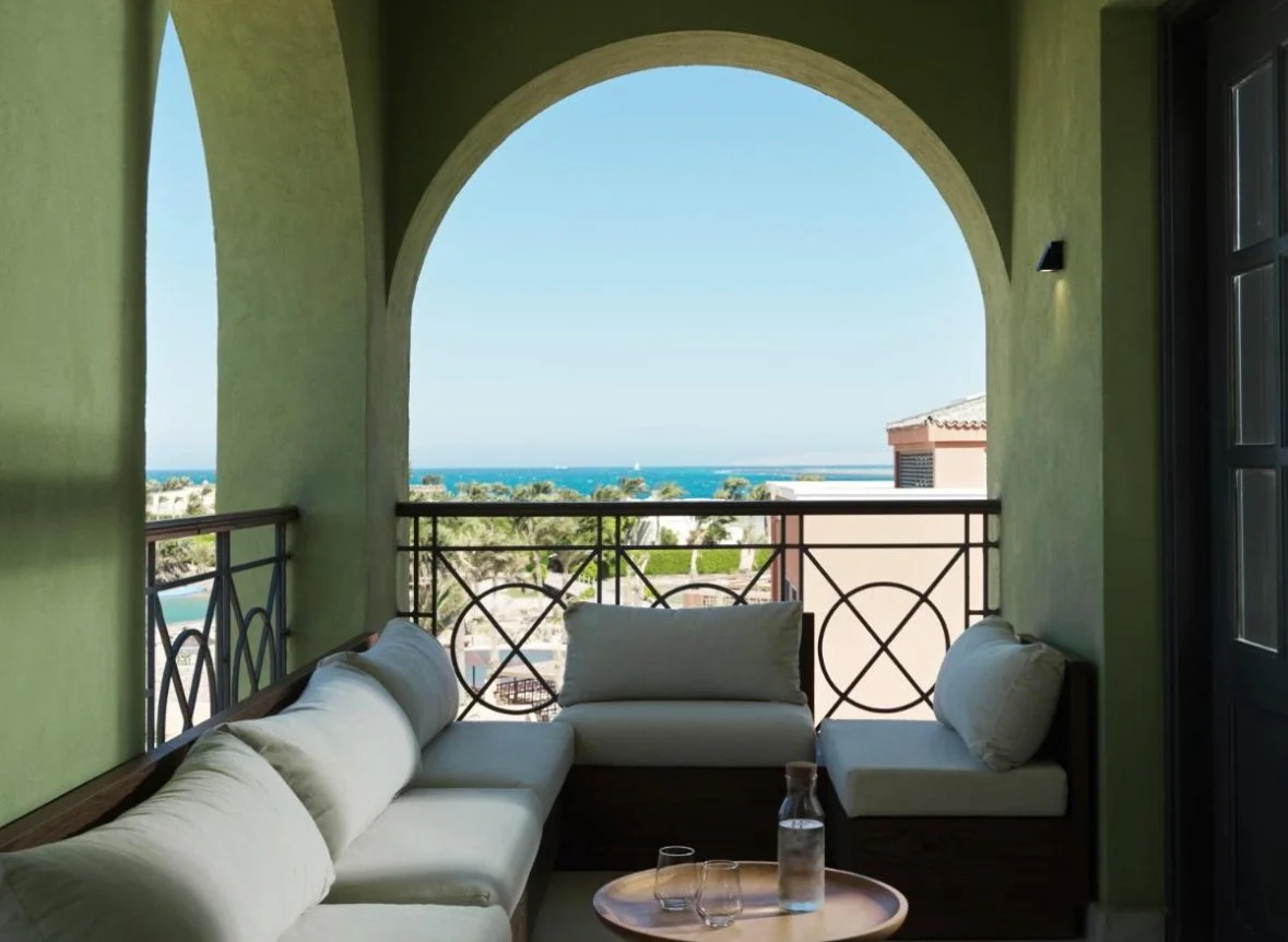 View of a balcony with a white outdoor sofa and a round wooden table holding a water bottle and glasses, overlooking a coastal scene with trees and the ocean in the distance.