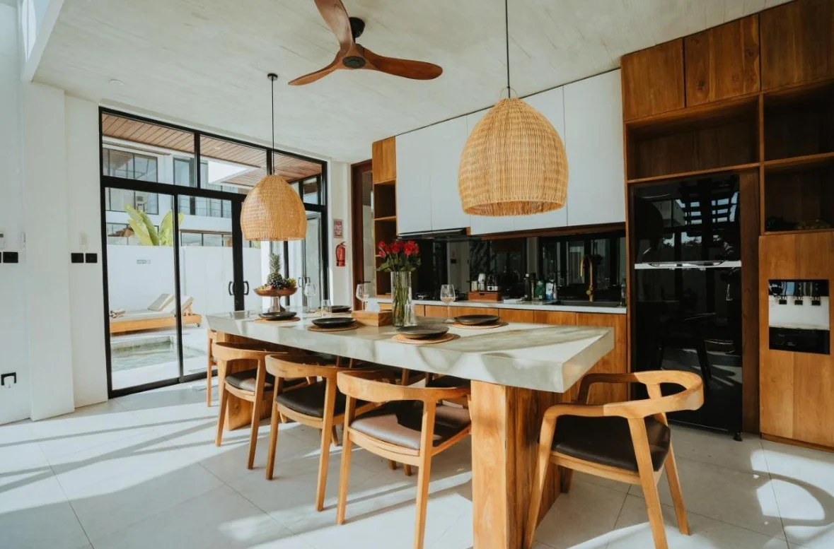 Modern kitchen with a marble island, six wooden chairs, woven pendant lights, wooden shelves, black appliances, and a sliding glass door leading outside.