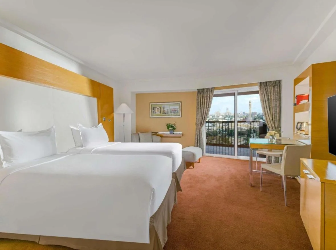 Hotel room with two queen beds, a sitting area with a small table and chair, a balcony with a view of a cityscape including a tall tower, a desk with a chair, and a television.