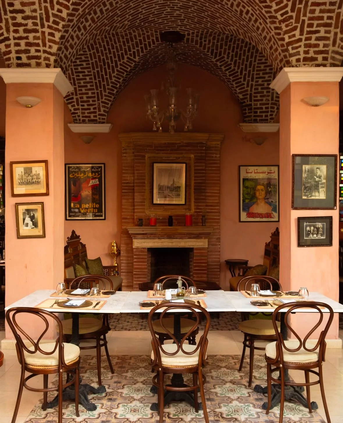 A dining area with a white marble table set for six people, surrounded by wooden chairs with upholstered seats. The room has pink walls adorned with vintage posters and black-and-white photographs. There is a brick fireplace with a framed picture abo