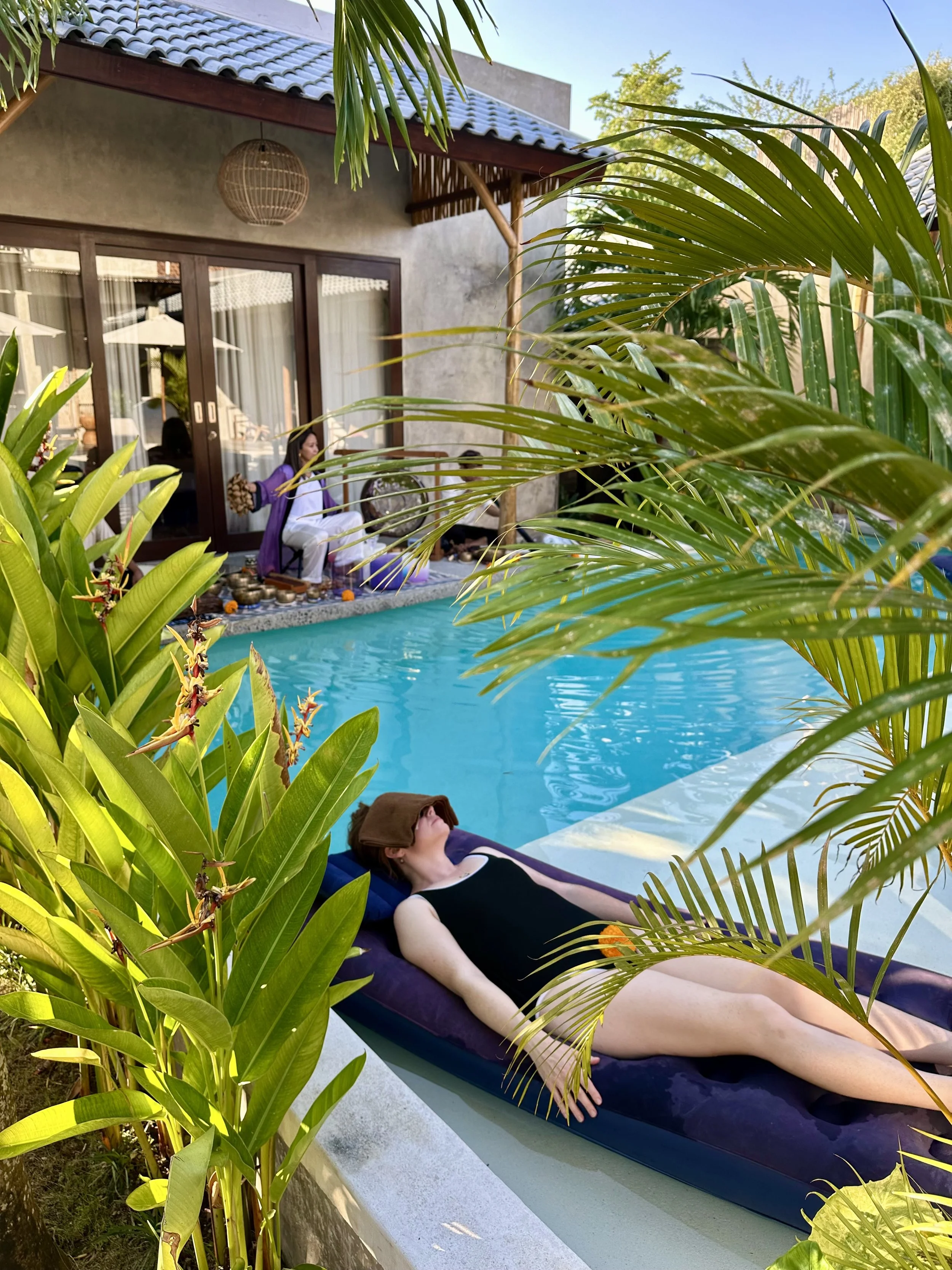 A woman relaxing on a pool float with her eyes closed and a towel over her face, surrounded by tropical plants at a backyard poolside area. Floating sound bath in Bali.