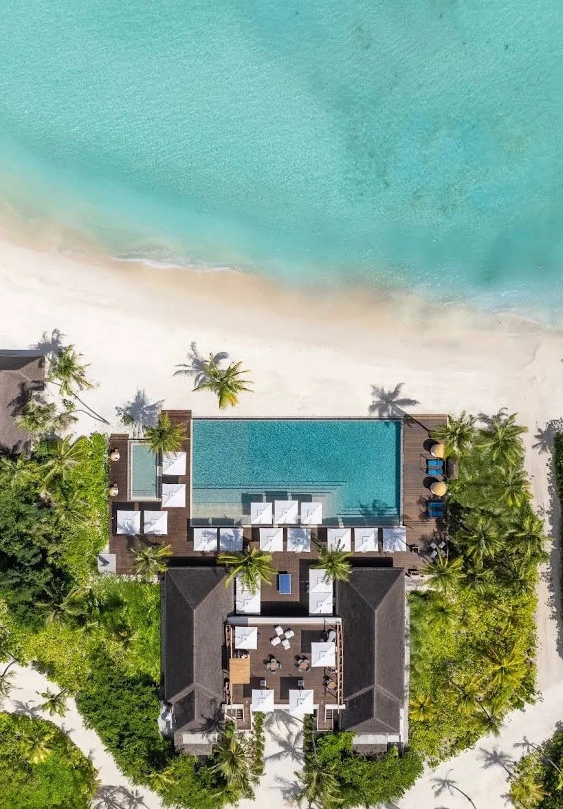 Aerial view of a beach resort in Bali with a large swimming pool, surrounded by palm trees, lounge chairs, and umbrellas, overlooking the ocean.
