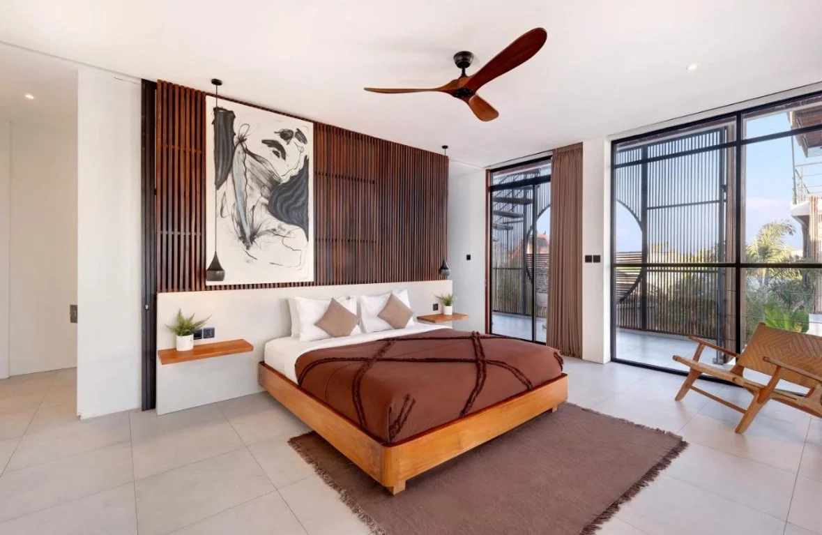 Modern bedroom with a large bed, brown blanket, white pillows, wooden side tables with plants, artwork on the wall, ceiling fan, and floor-to-ceiling windows with balcony and view outside.