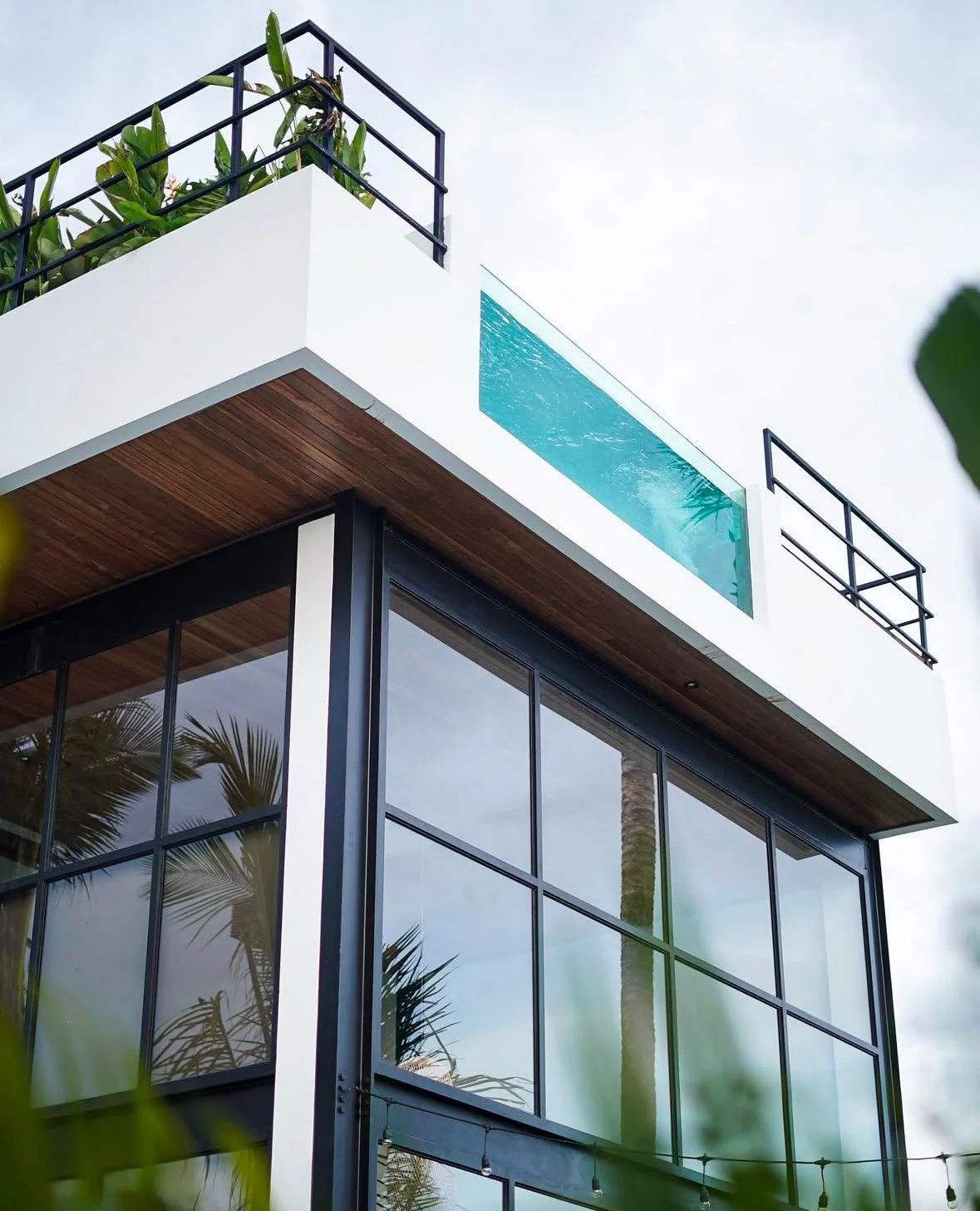 Modern multi-story house with large glass windows, white walls, wooden accents, a rooftop pool with clear blue water, and greenery including palm trees.