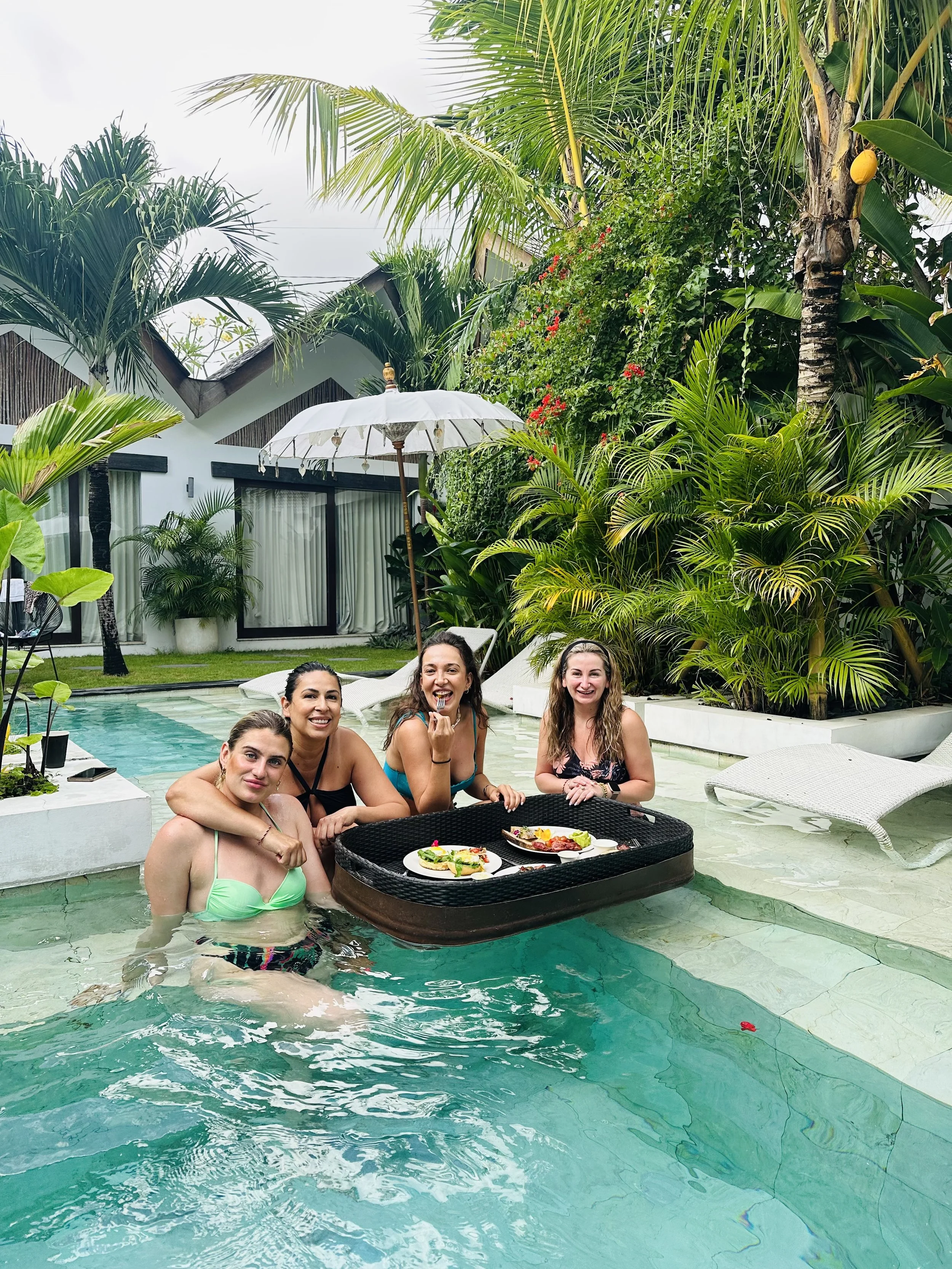 Four women enjoying a poolside meal, with lush tropical plants, lounge chairs, and a white umbrella in the background in Canggu, Bali.