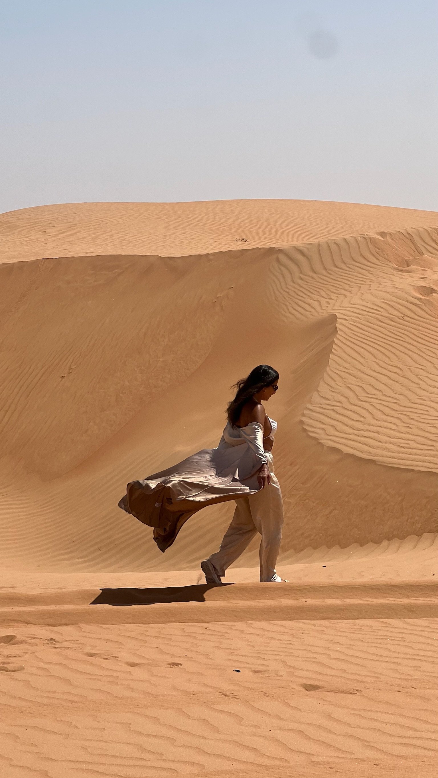A woman in beige clothing walking across sand dunes in a a desert in Egypt.