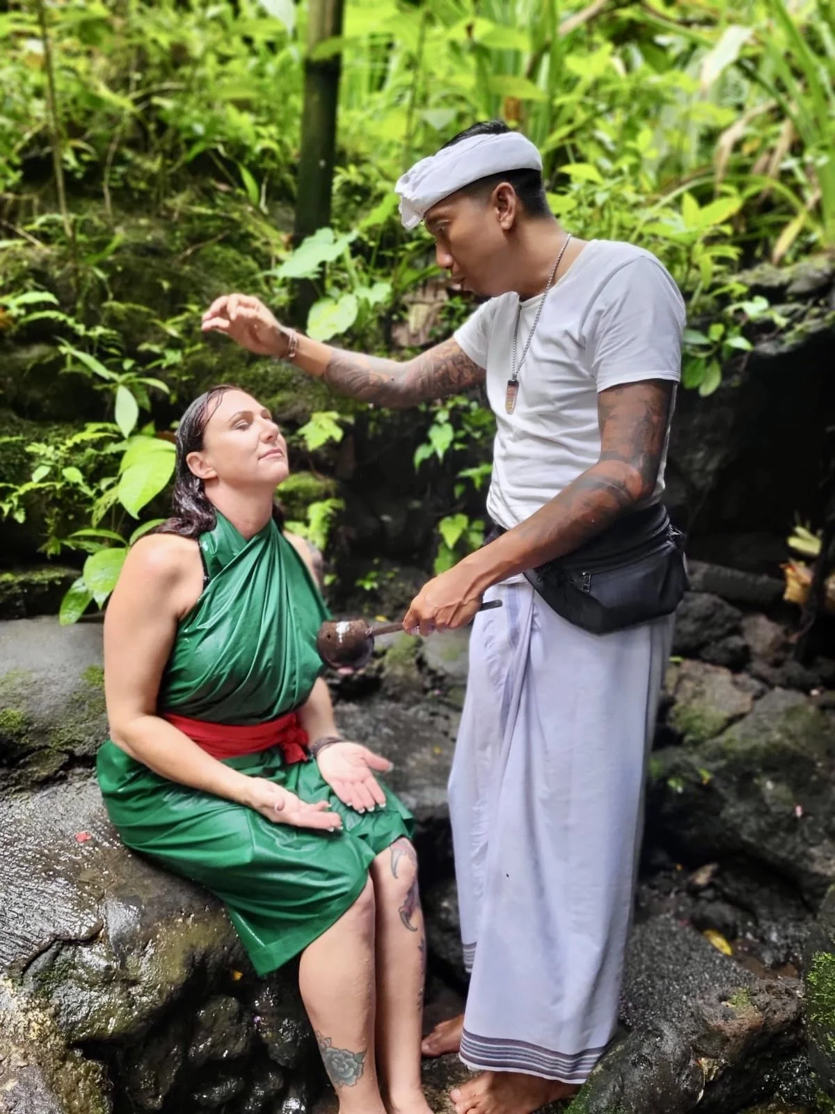 A woman dressed in a green and red outfit sitting in a natural, lush jungle setting, receiving a spiritual blessing from a man wearing traditional white and gray clothing and tattoos, with dense jungle foliage in the background in Bali.