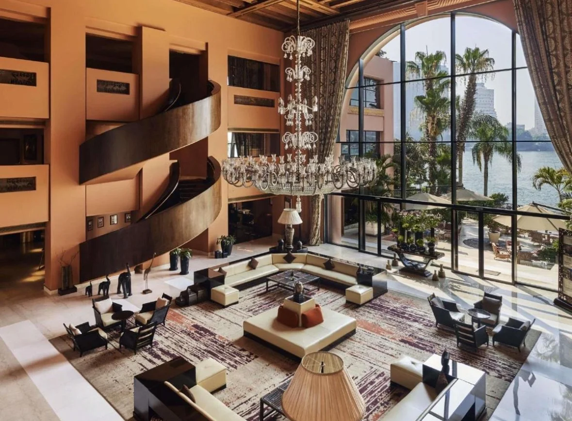 Spacious hotel lobby with large floor-to-ceiling windows overlooking a river with palm trees. The room has a chandelier, a large sectional sofa, several armchairs, and decorative elements like plants and sculptures.