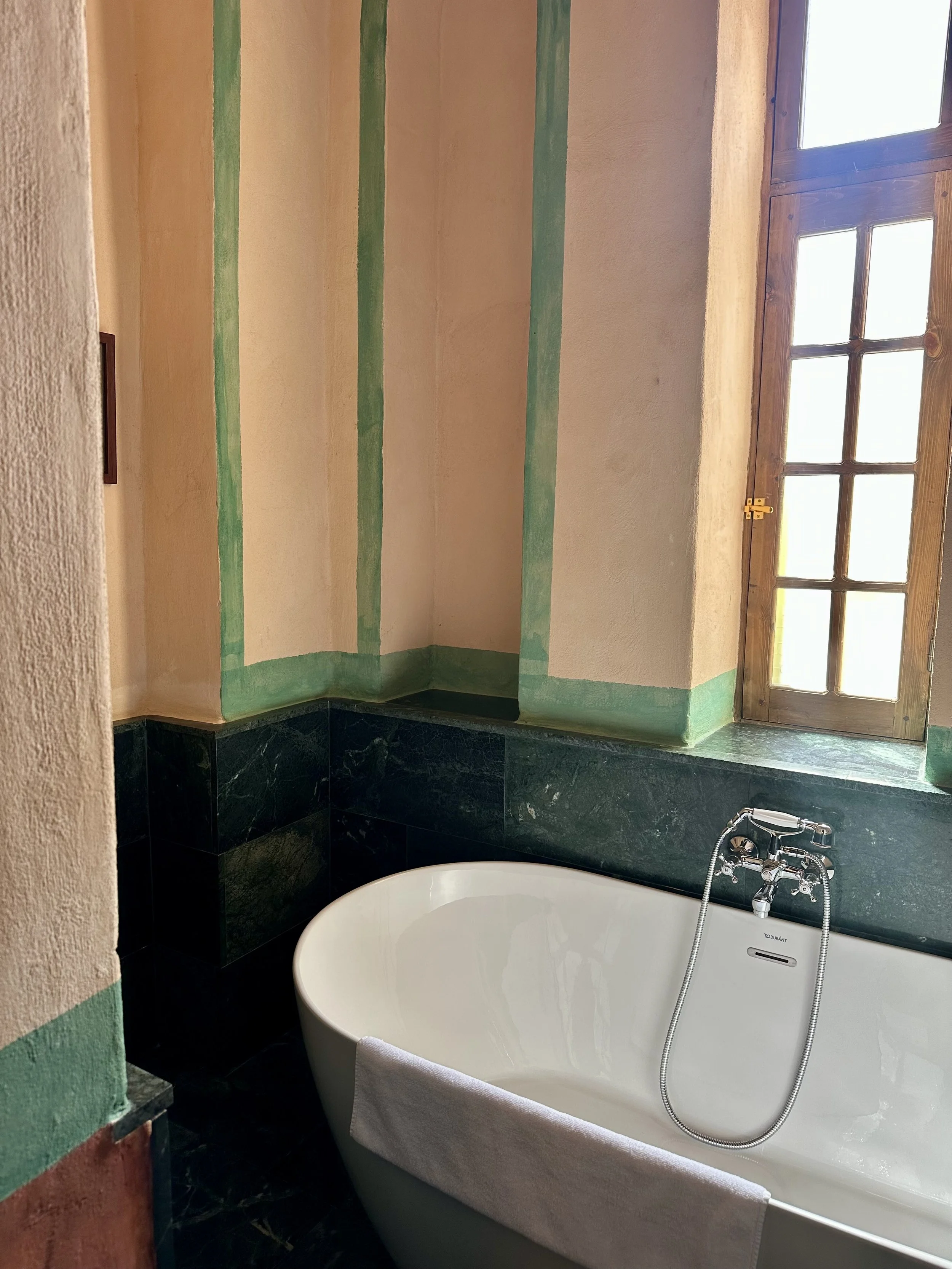 Clawfoot bathtub with a towel draped over the side, in a bathroom with a window, wooden window frame, green stone tiles, and painted walls with green accents.