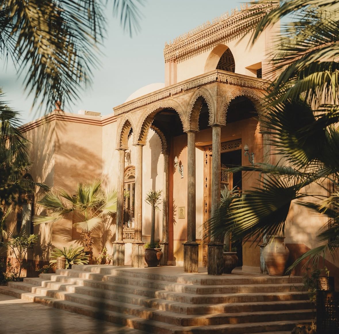 An ornate building with architectural arches and columns, surrounded by lush palm trees and potted plants, with steps leading up to the entrance, bathed in warm sunlight.