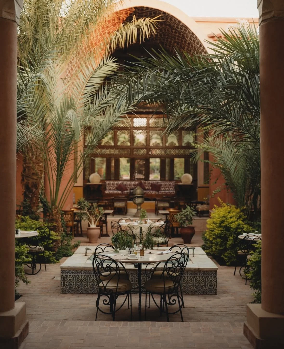 Elegant outdoor dining area with wrought iron tables and chairs, lush green plants, and a Moroccan-style interior with arched windows and intricate woodwork in the background.