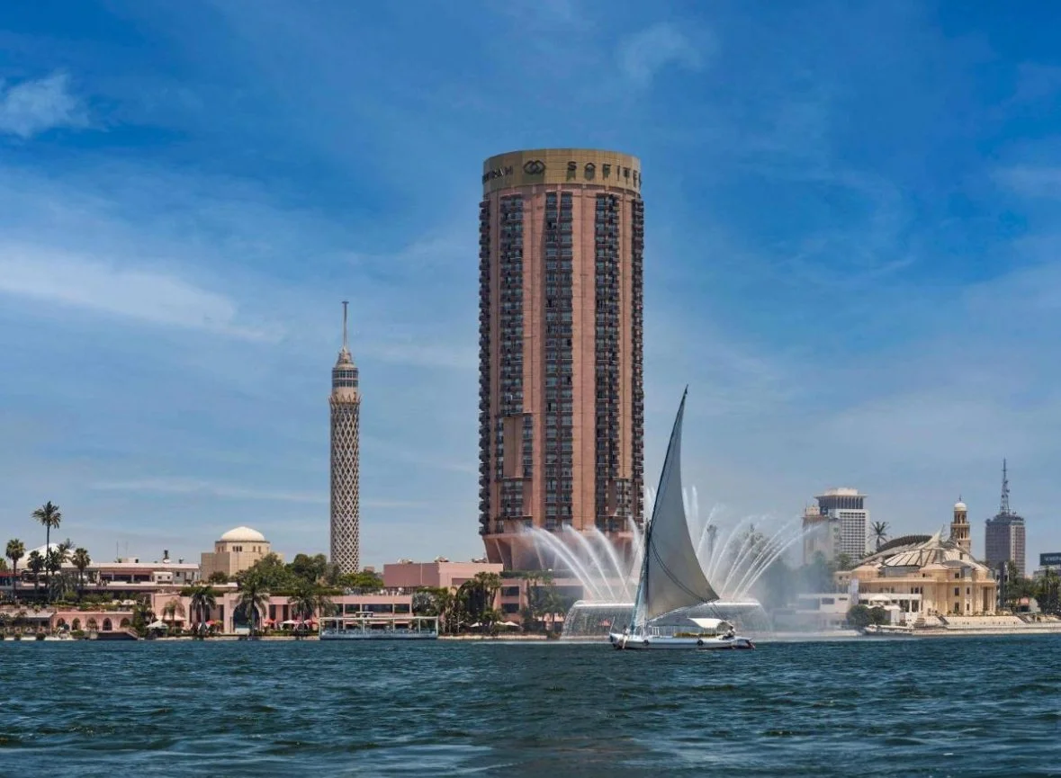 Cityscape of a waterfront with high-rise buildings, a sailboat, and a fountain on the water with streams shooting upward. Blue sky with some clouds.