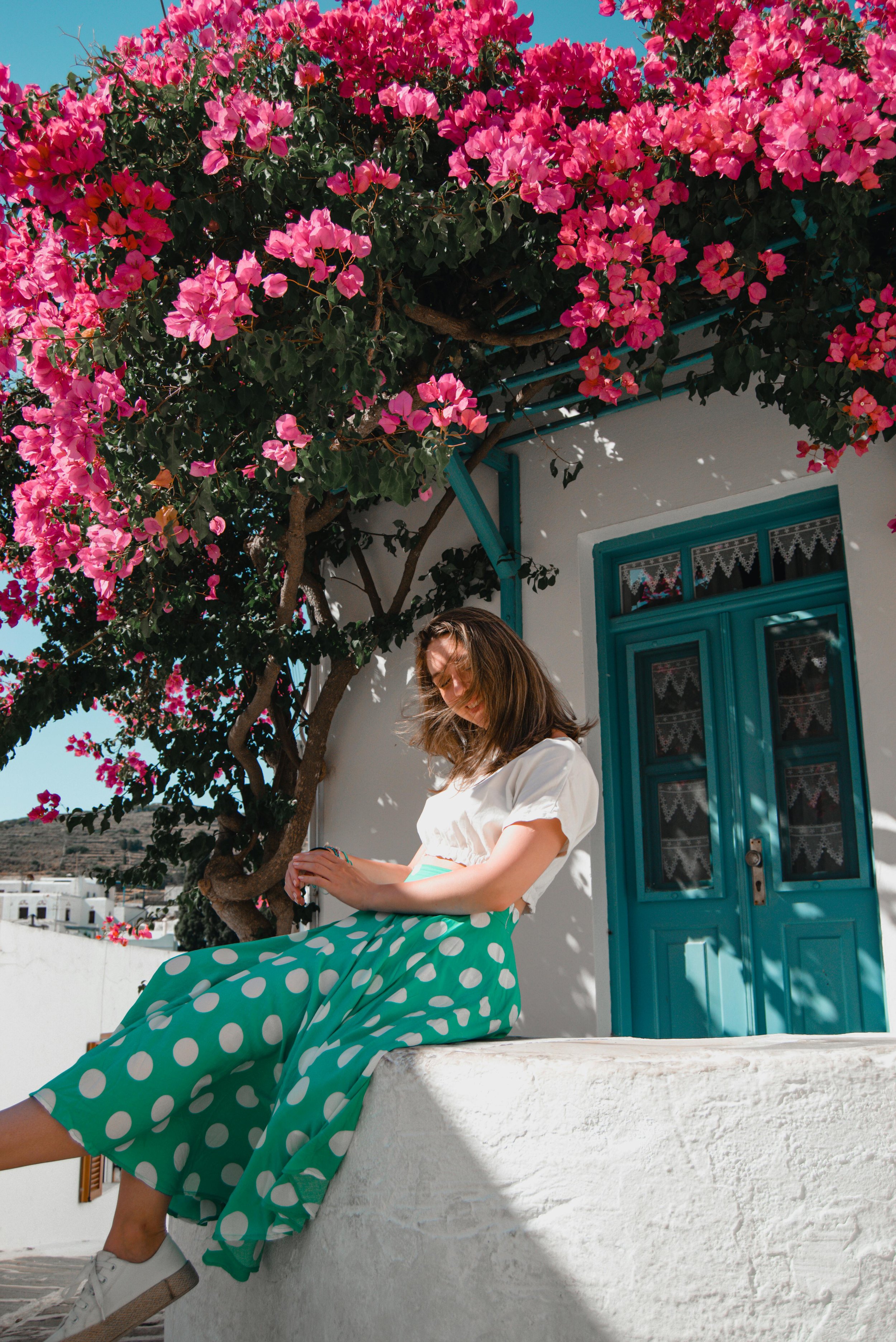 A woman sitting on a white wall in front of a white building with a turquoise door, under a large pink bougainvillea tree, wearing white top, green polka dot skirt, and white shoes in Kano twin villages in Bali.