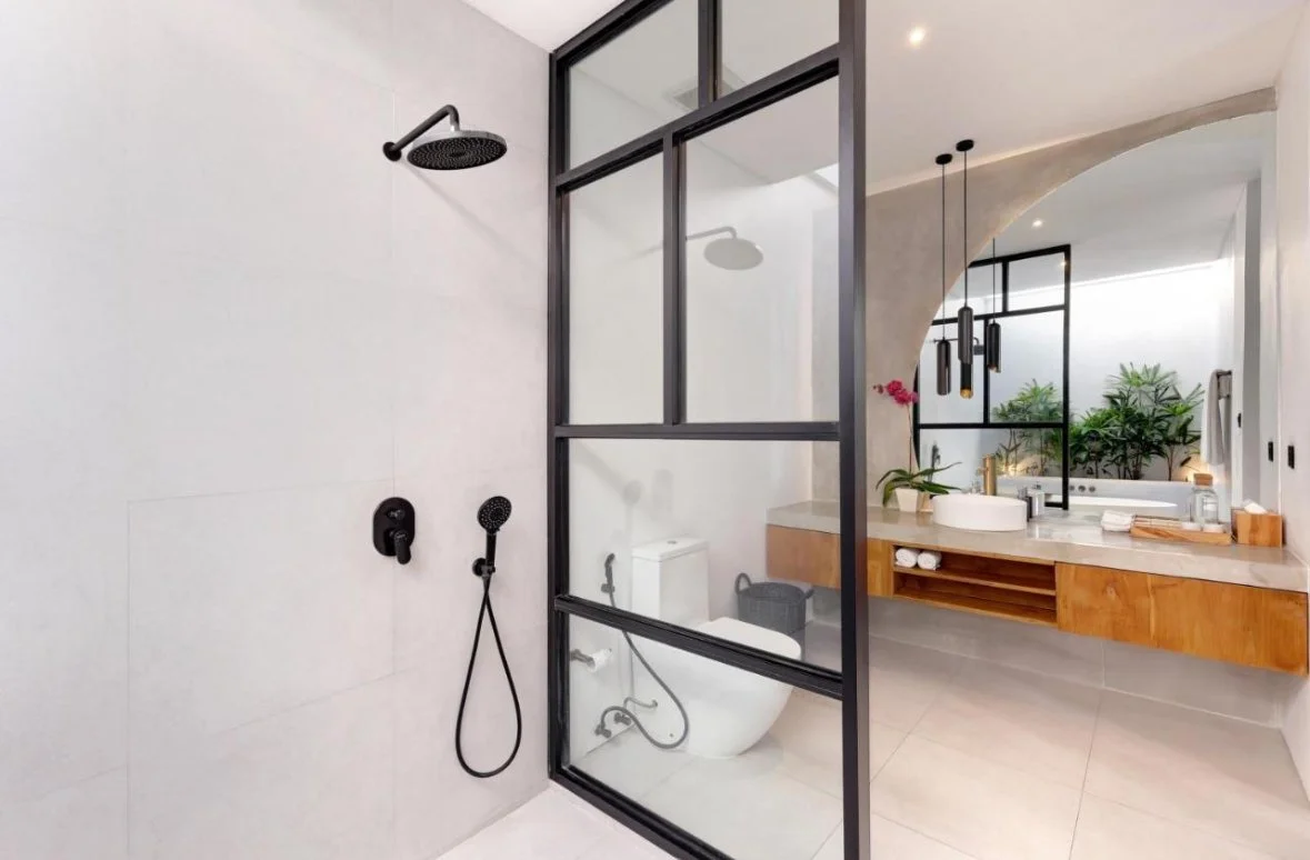 Modern bathroom featuring a walk-in shower with black fixtures, a glass partition, a floating wooden vanity with a white vessel sink, and green plants in the background.