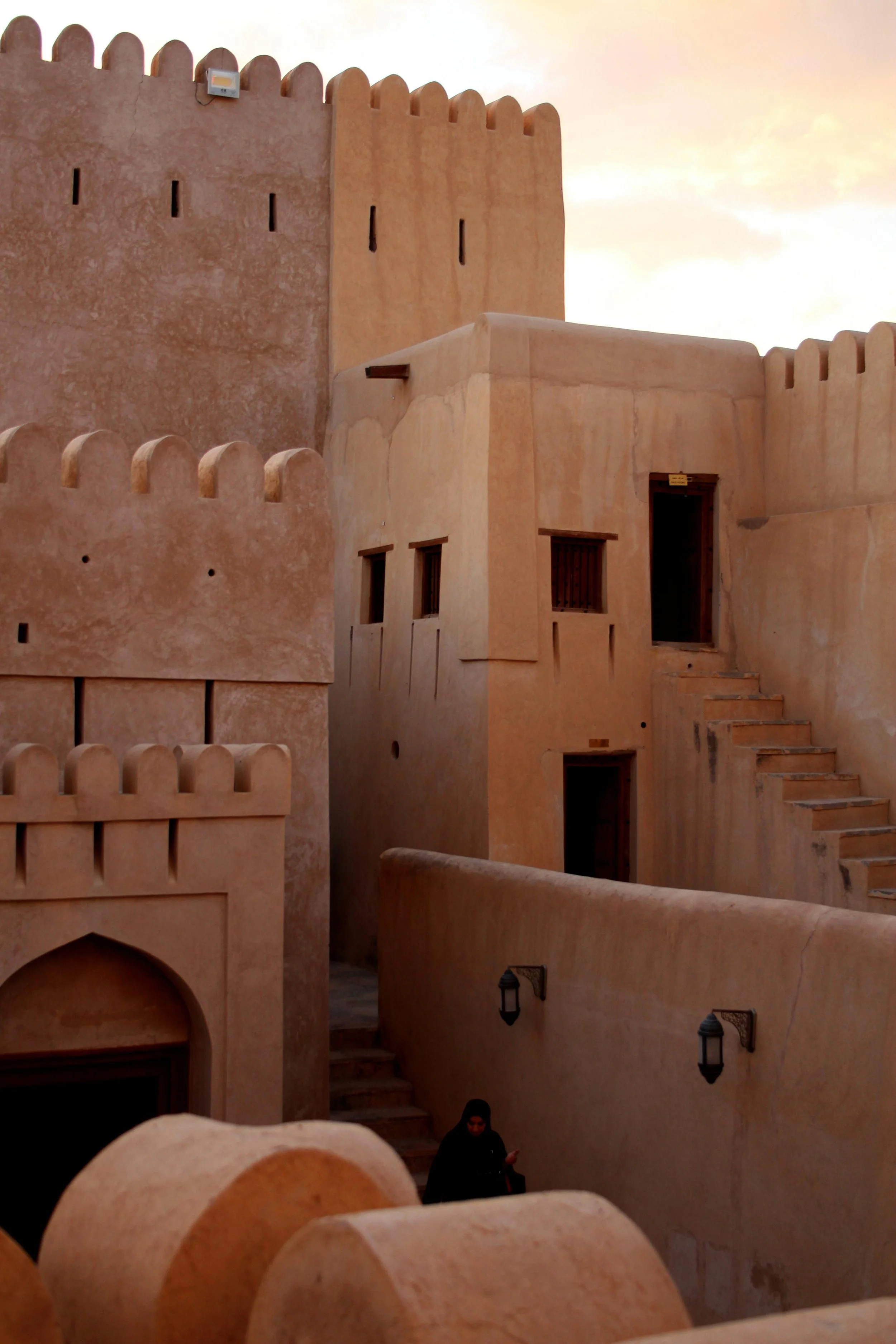 A person in black clothing standing near tan, traditional desert-style adobe buildings with stepped stairs and wooden windows.
