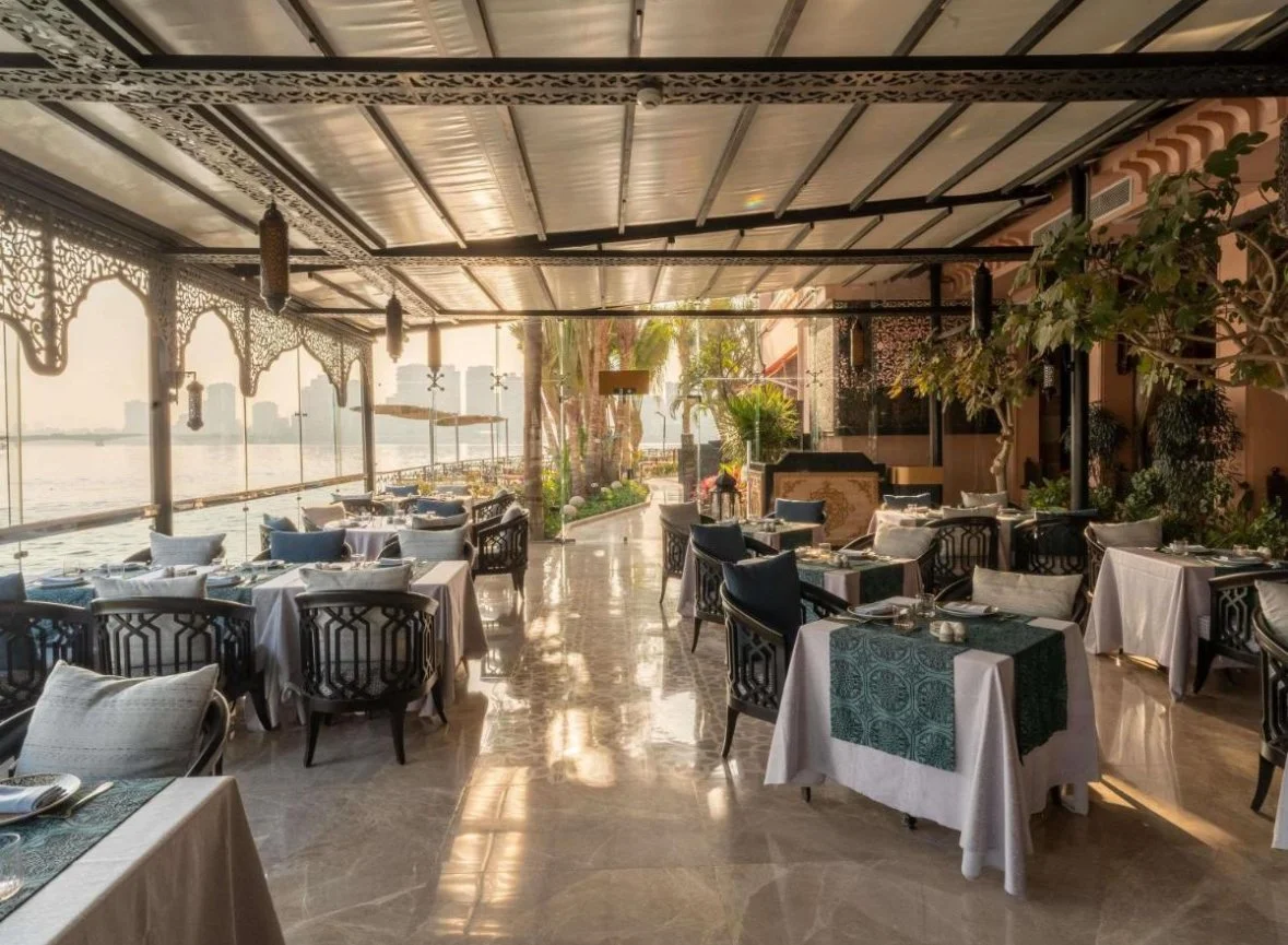 An elegant restaurant overlooking a body of water with outdoor seating, white tablecloths, and decorative chairs, featuring lush tropical plants and sunset lighting.