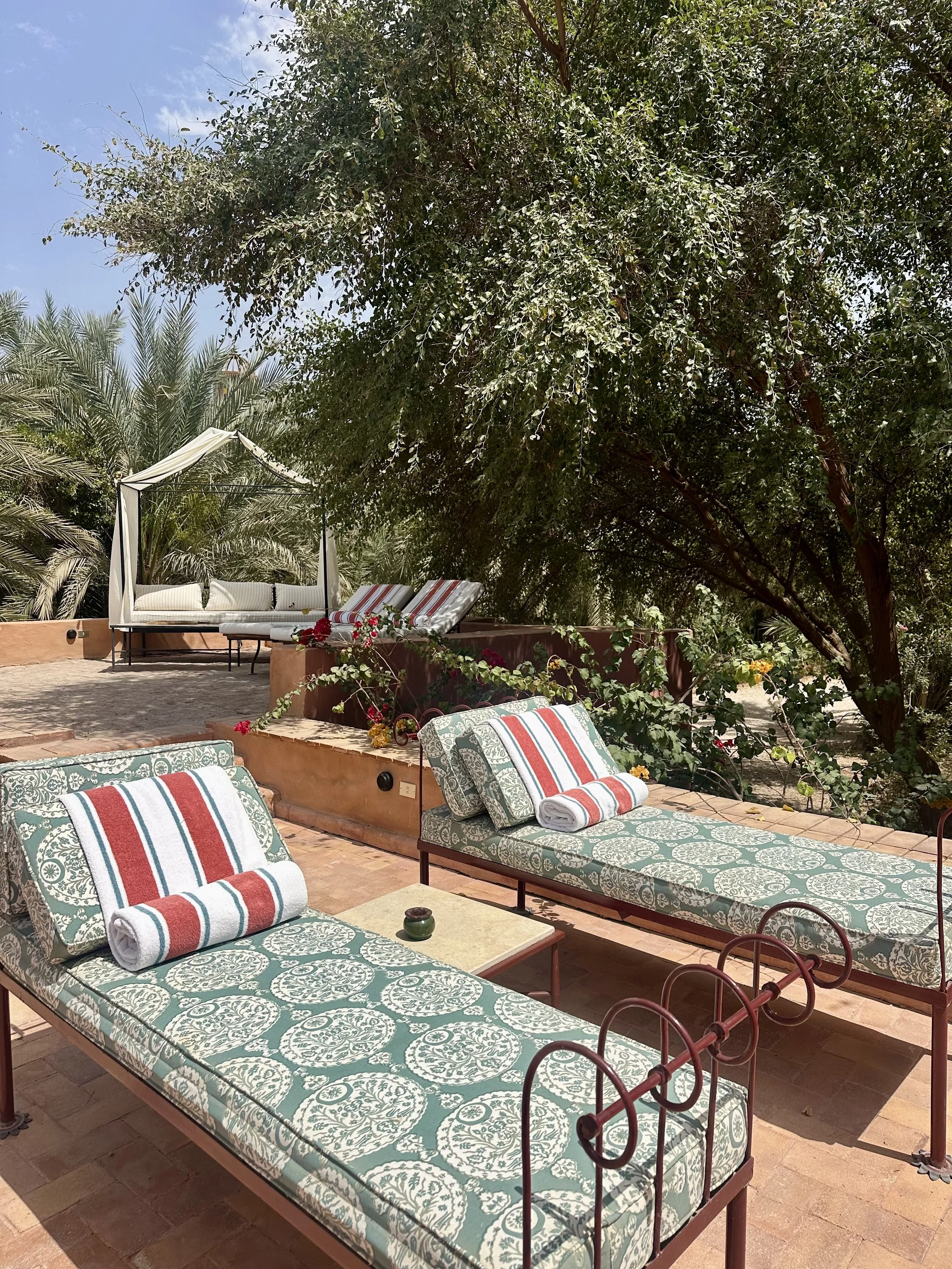 Outdoor patio with two lounge chairs featuring patterned cushions and pillows, a small table with a candle, lush green trees, and a white canopy bed with striped cushions in the background.
