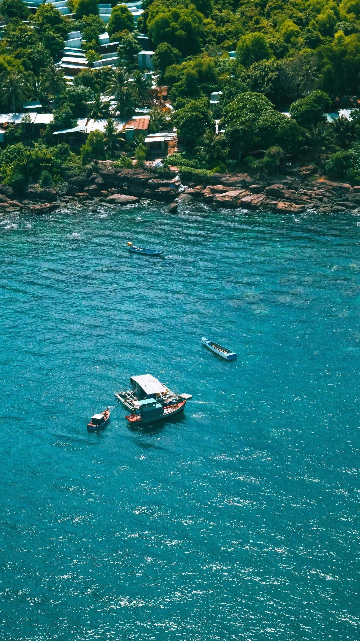 Boats anchored in clear blue water near a rocky shoreline, with a lush green hillside and small houses in the background in Bali.